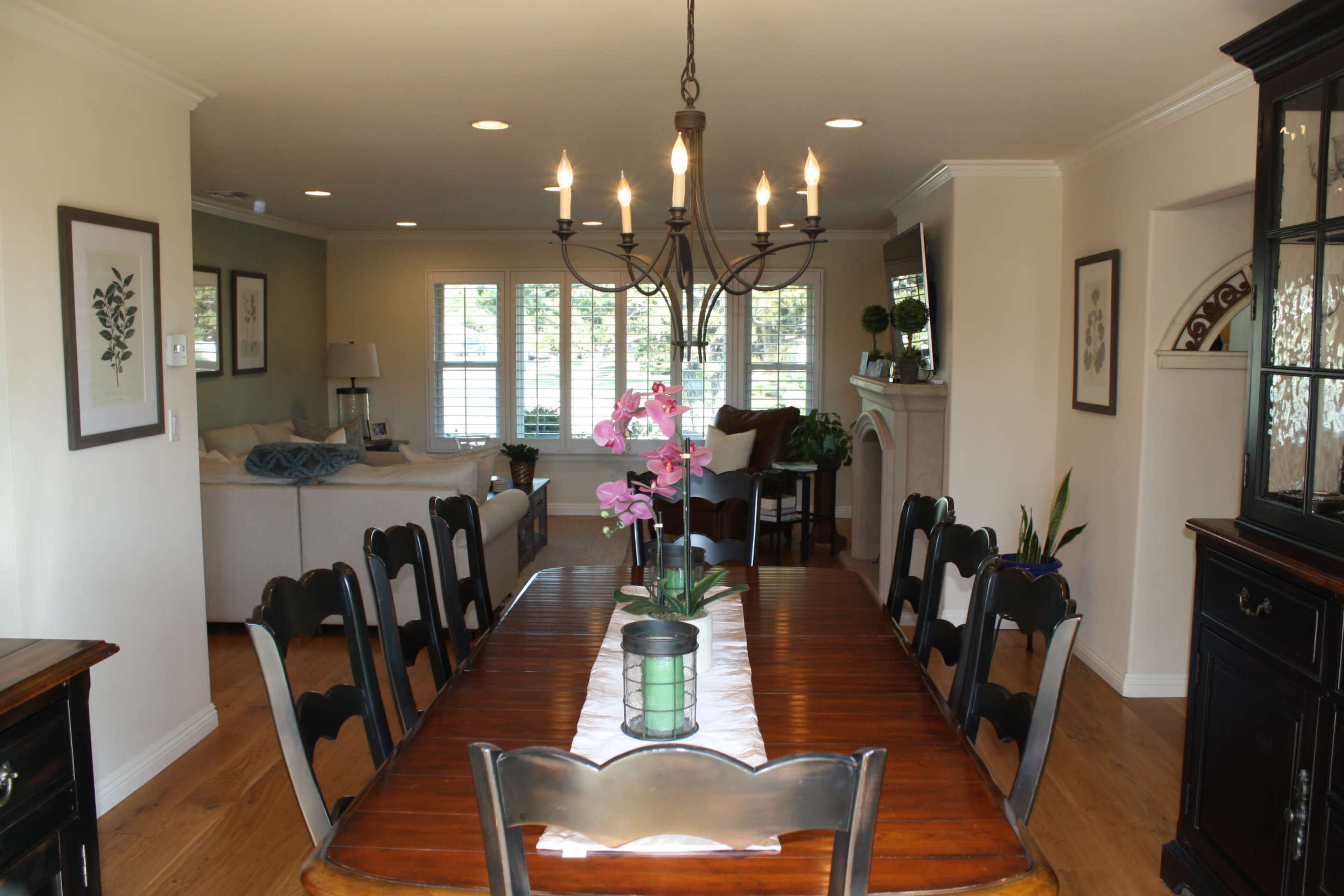 A long wooden dining table with black chairs is centered under a chandelier, flanked by light-colored walls and a view into a living area with a couch.