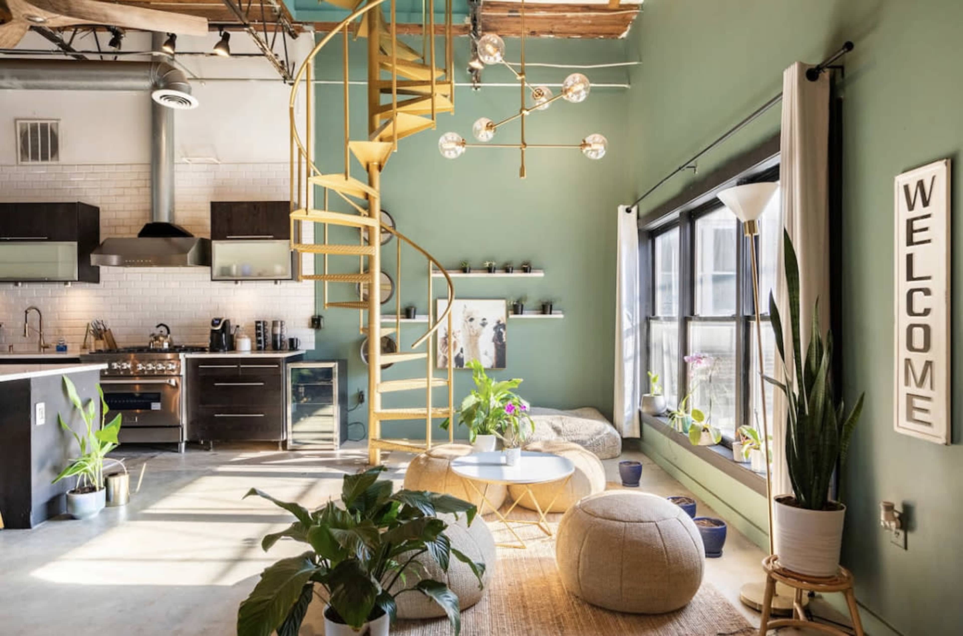 A modern living space featuring a spiral staircase, green walls, and a mix of plants and furniture arranged around a central area with a welcome sign.