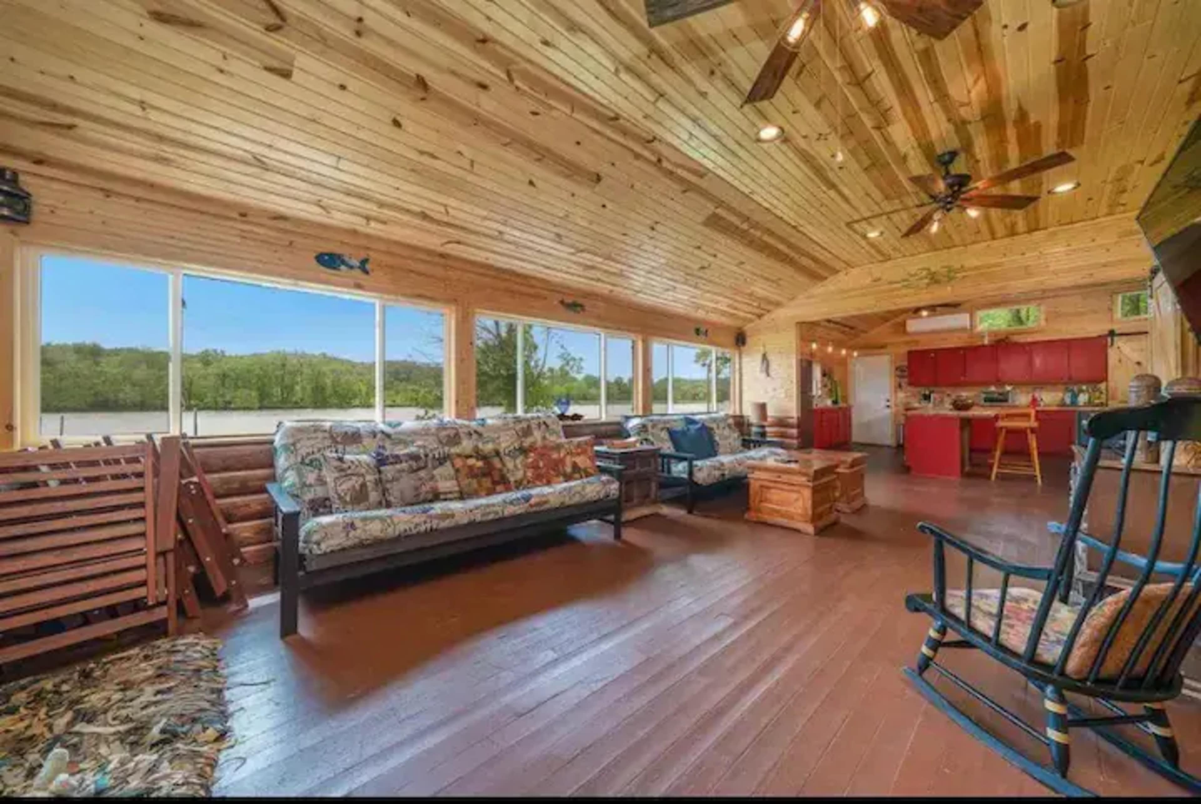 A spacious, wooden-paneled living area with large windows, comfortable seating, and a kitchen area in the background.