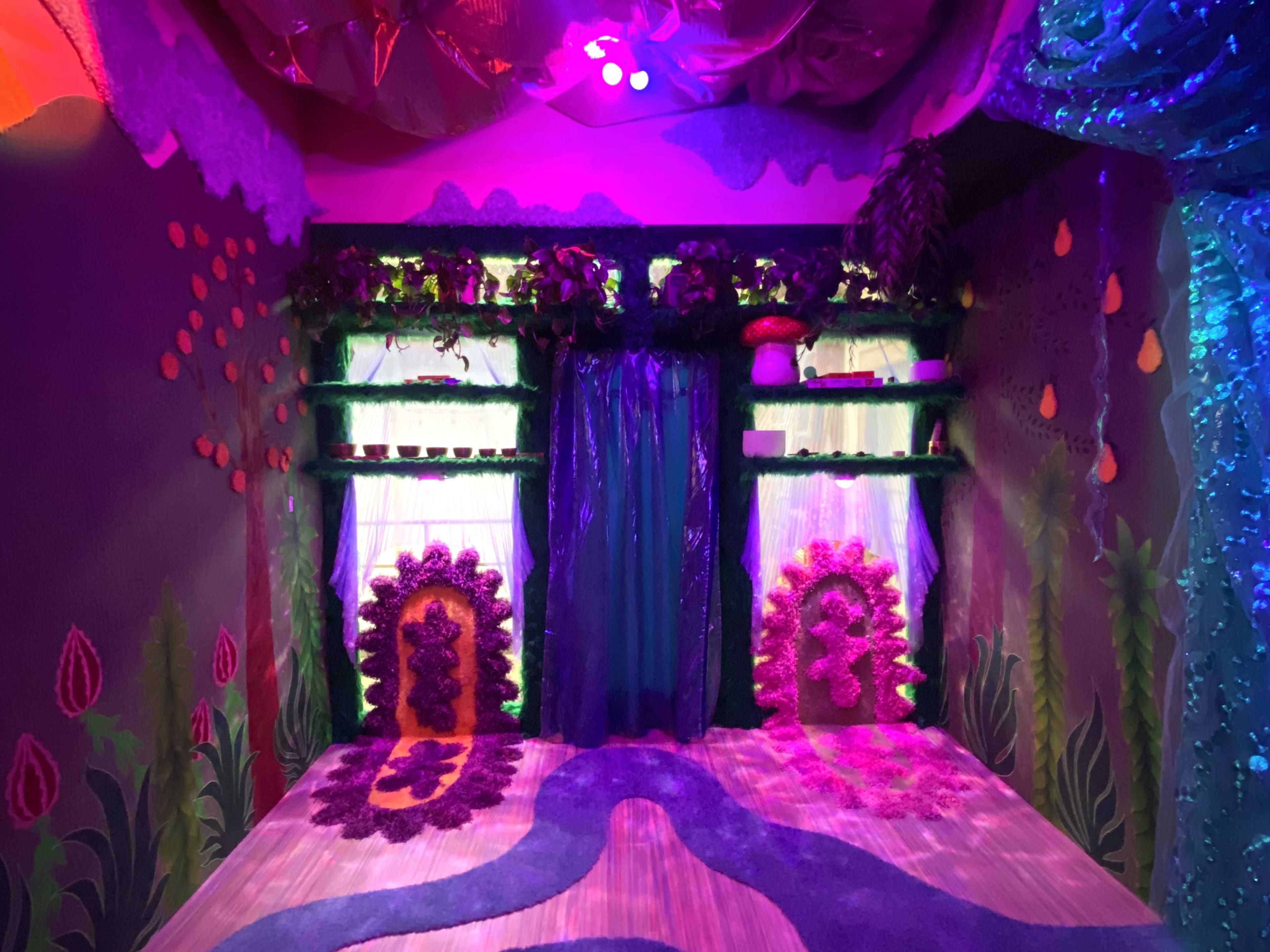 The image shows a brightly decorated room with colorful walls, two windows draped in sheer fabric, and a whimsical patterned floor.