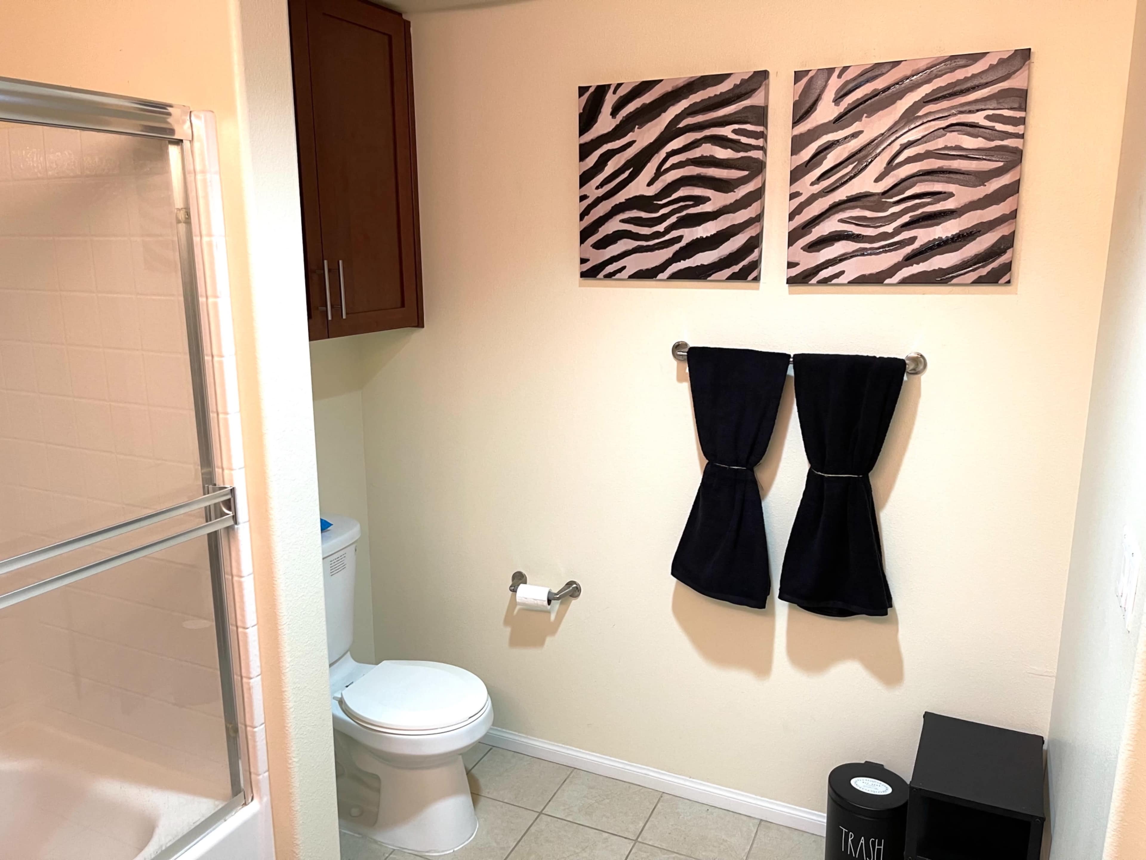 A bathroom with a shower, a toilet, two black towels hanging on a towel rack, and two zebra-patterned paintings on the wall.