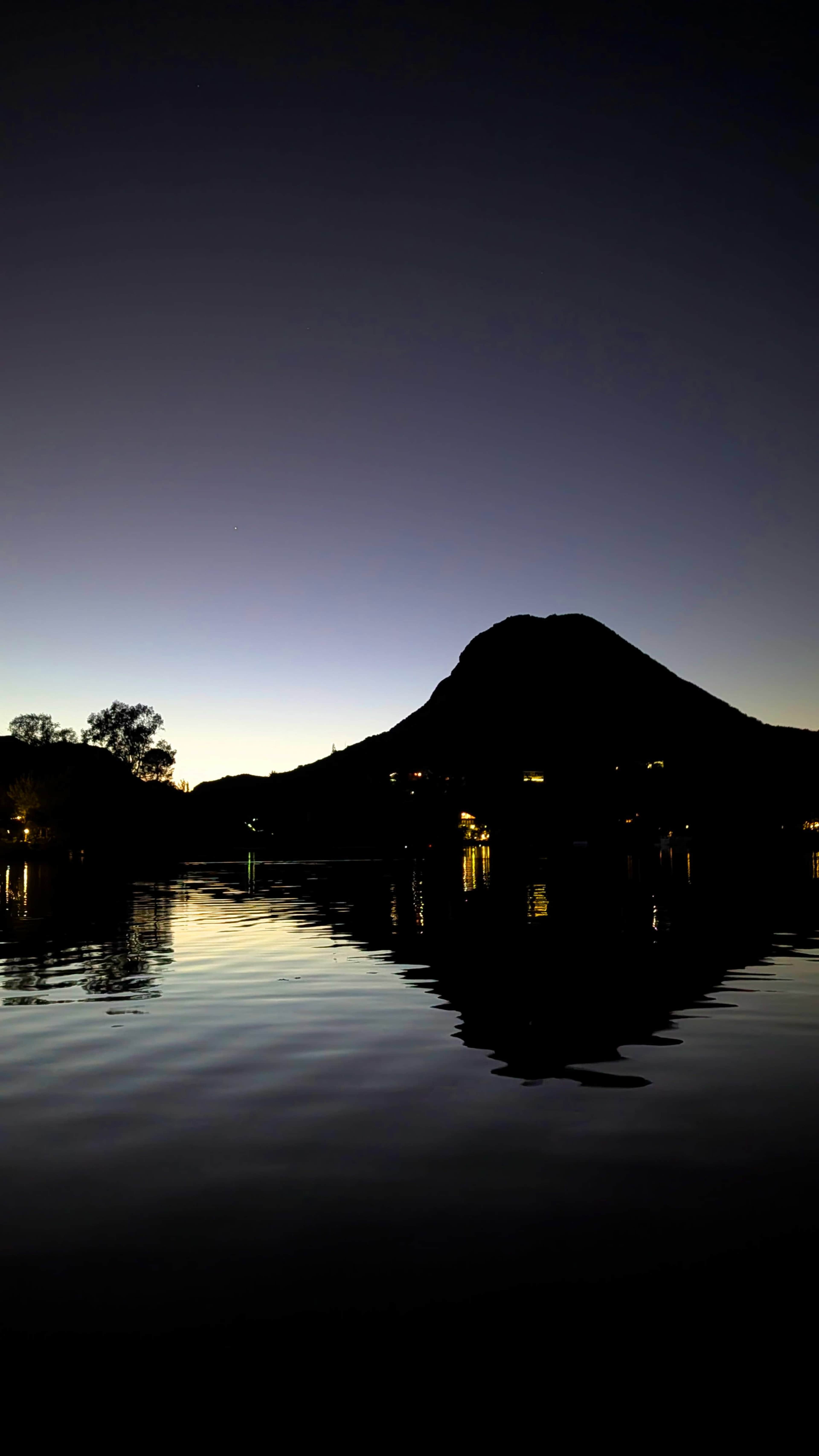 A mountain silhouette rises against a twilight sky, reflected in the calm water of a lake with faint lights from nearby buildings.