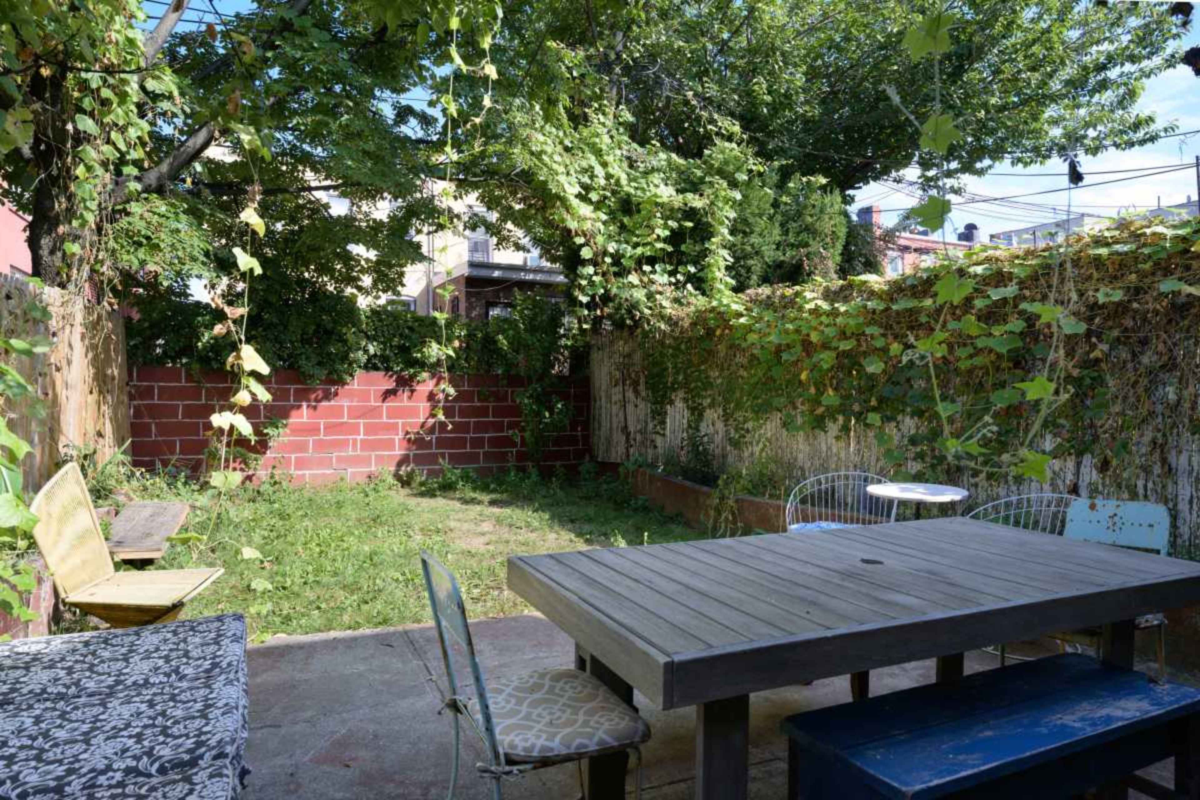 The image shows an overgrown backyard with a wooden table and chairs surrounded by green foliage and a brick wall.
