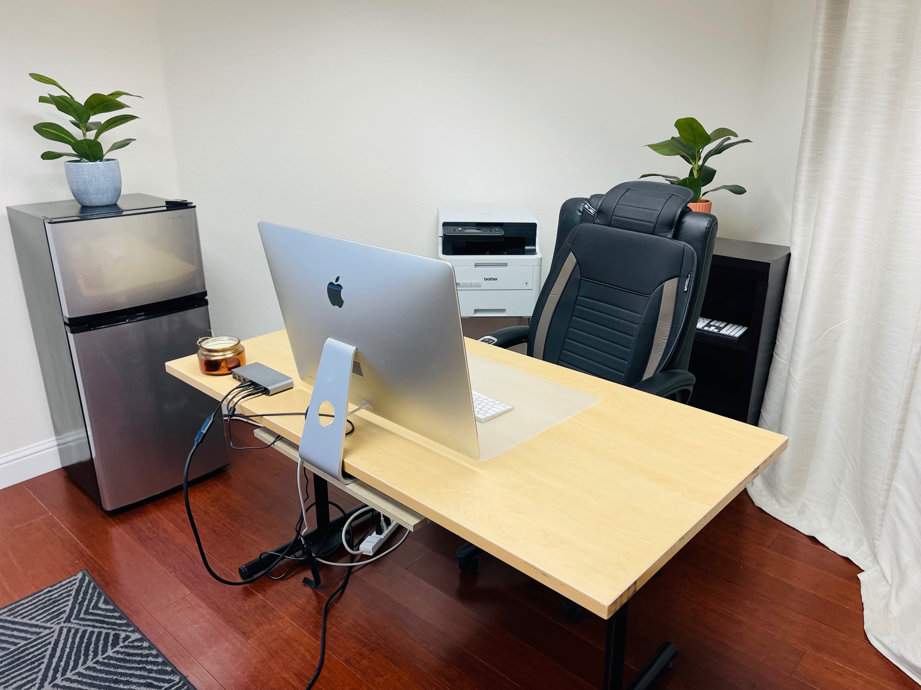 The image shows a modern office setup featuring a wooden desk with an iMac, a black ergonomic chair, a small fridge, a printer, and potted plants.