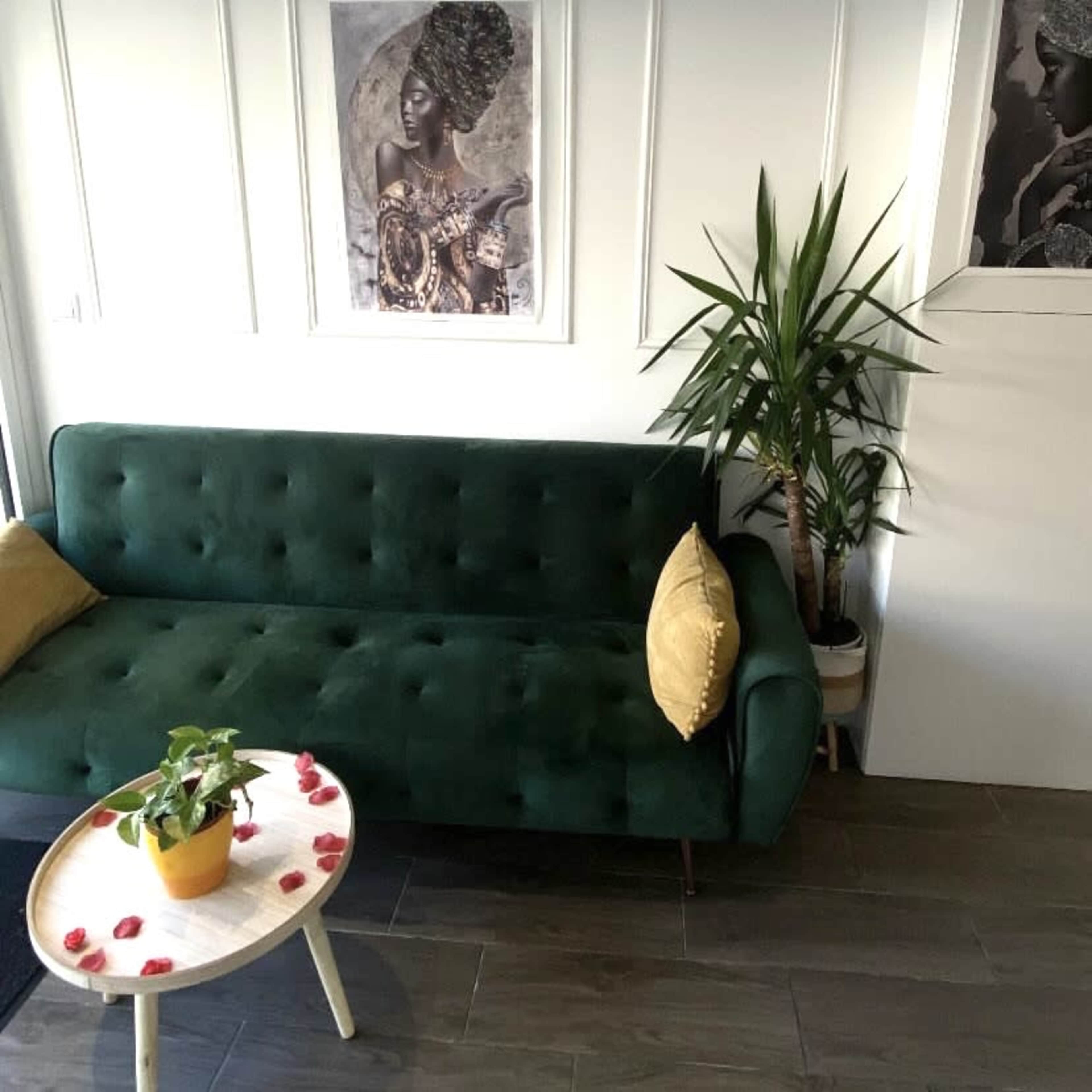 A green sofa with a yellow pillow and a small round table adorned with flower petals, alongside a potted plant against a white wall featuring framed artwork.
