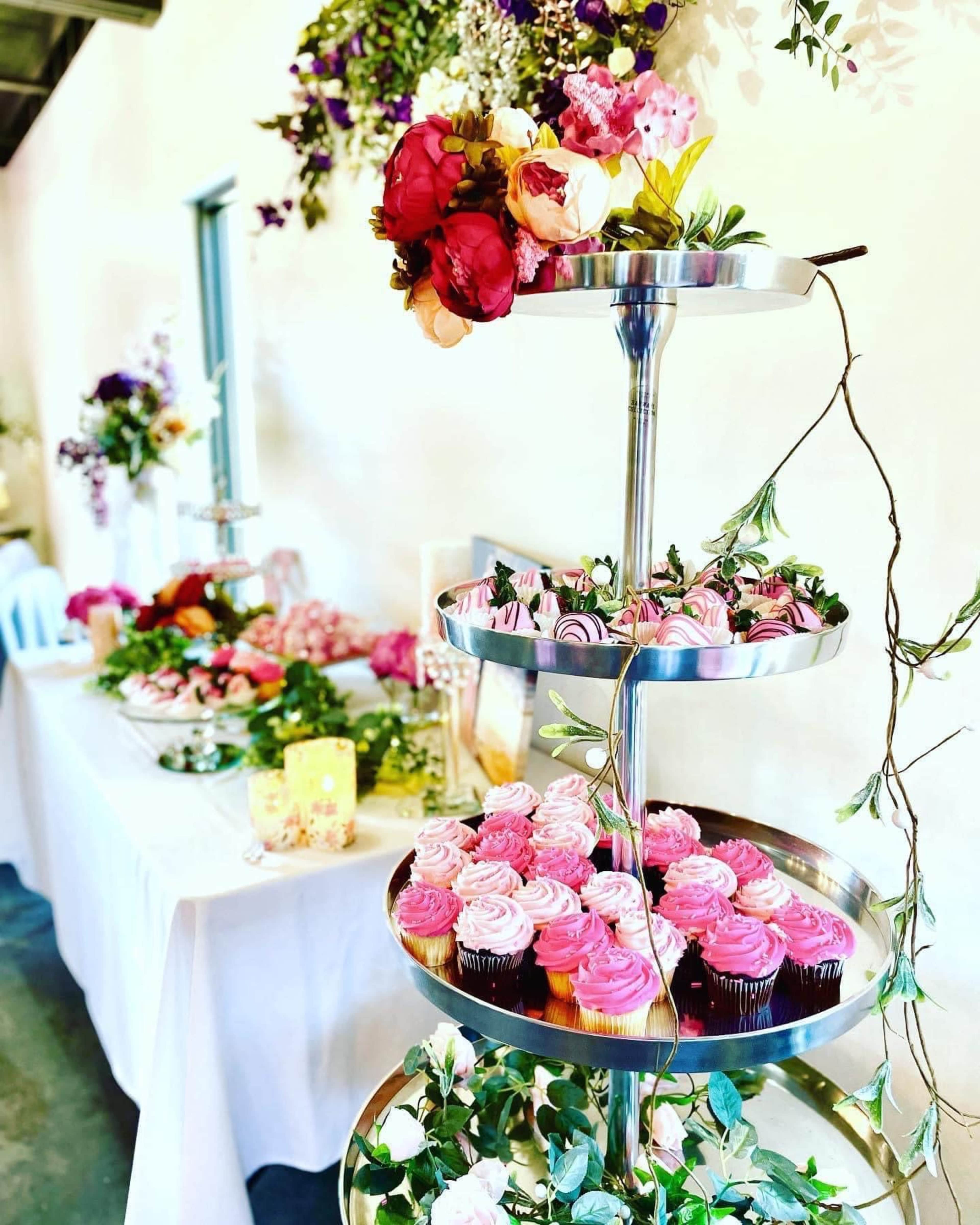 A multi-tiered stand displays an array of pink and purple cupcakes surrounded by floral decorations in a bright setting.