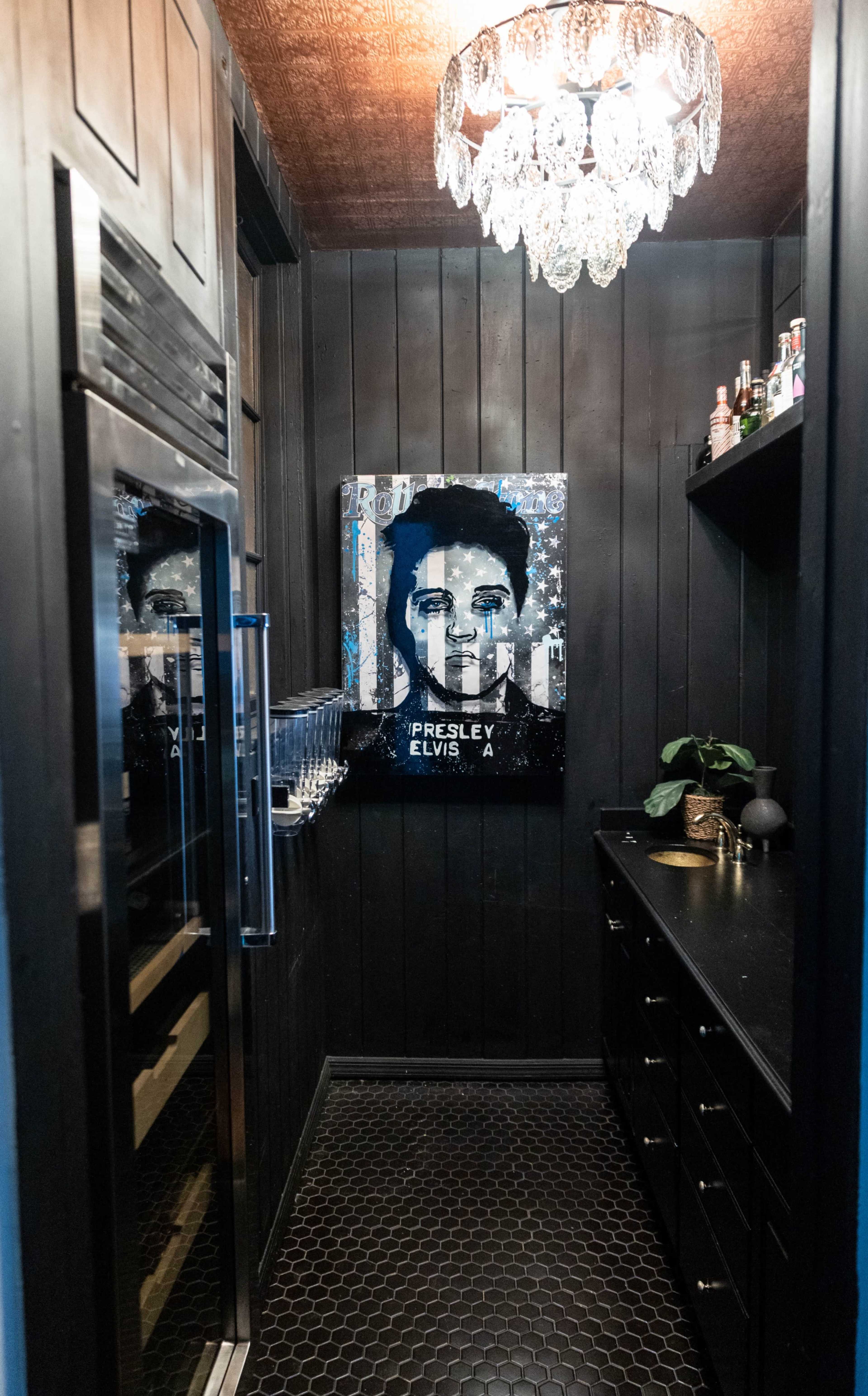 The image shows a dark room with a contemporary design featuring a refrigerator, a counter with bottles, and a large portrait of Elvis Presley on the wall.