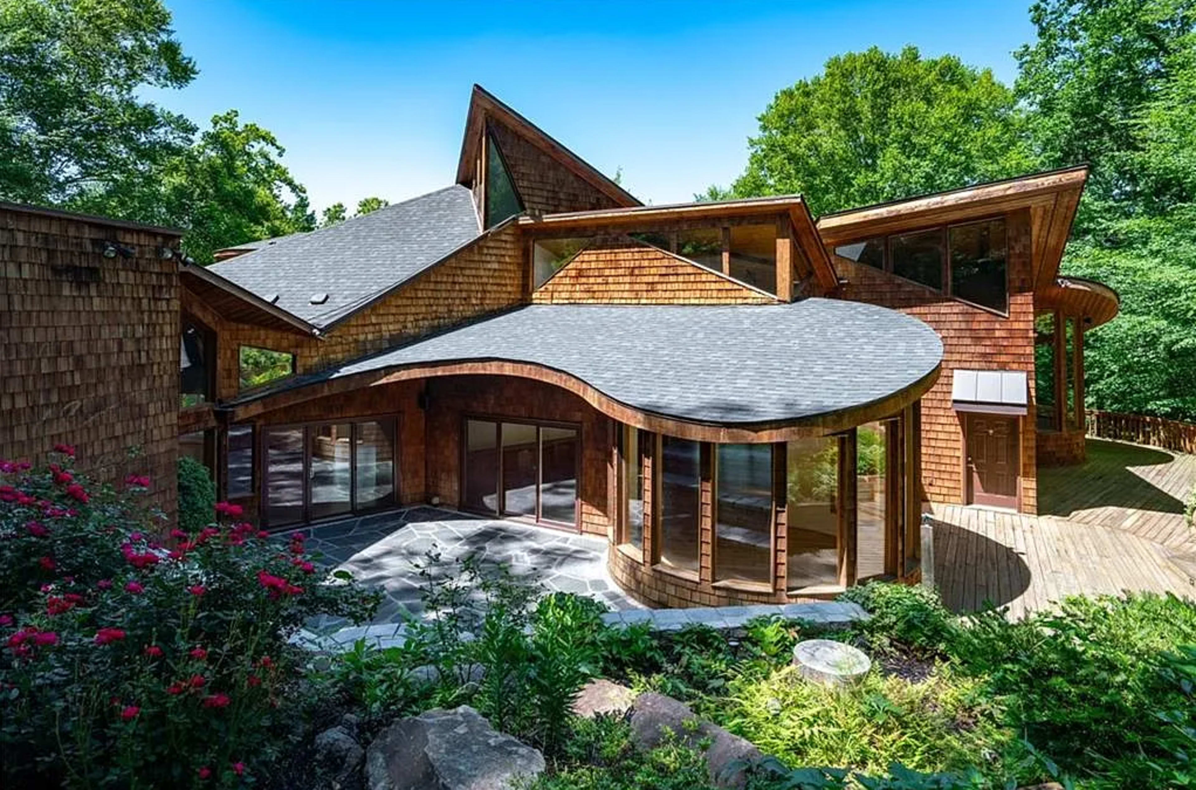 A modern wooden house with a curved roof and large glass windows is surrounded by greenery and flowers.