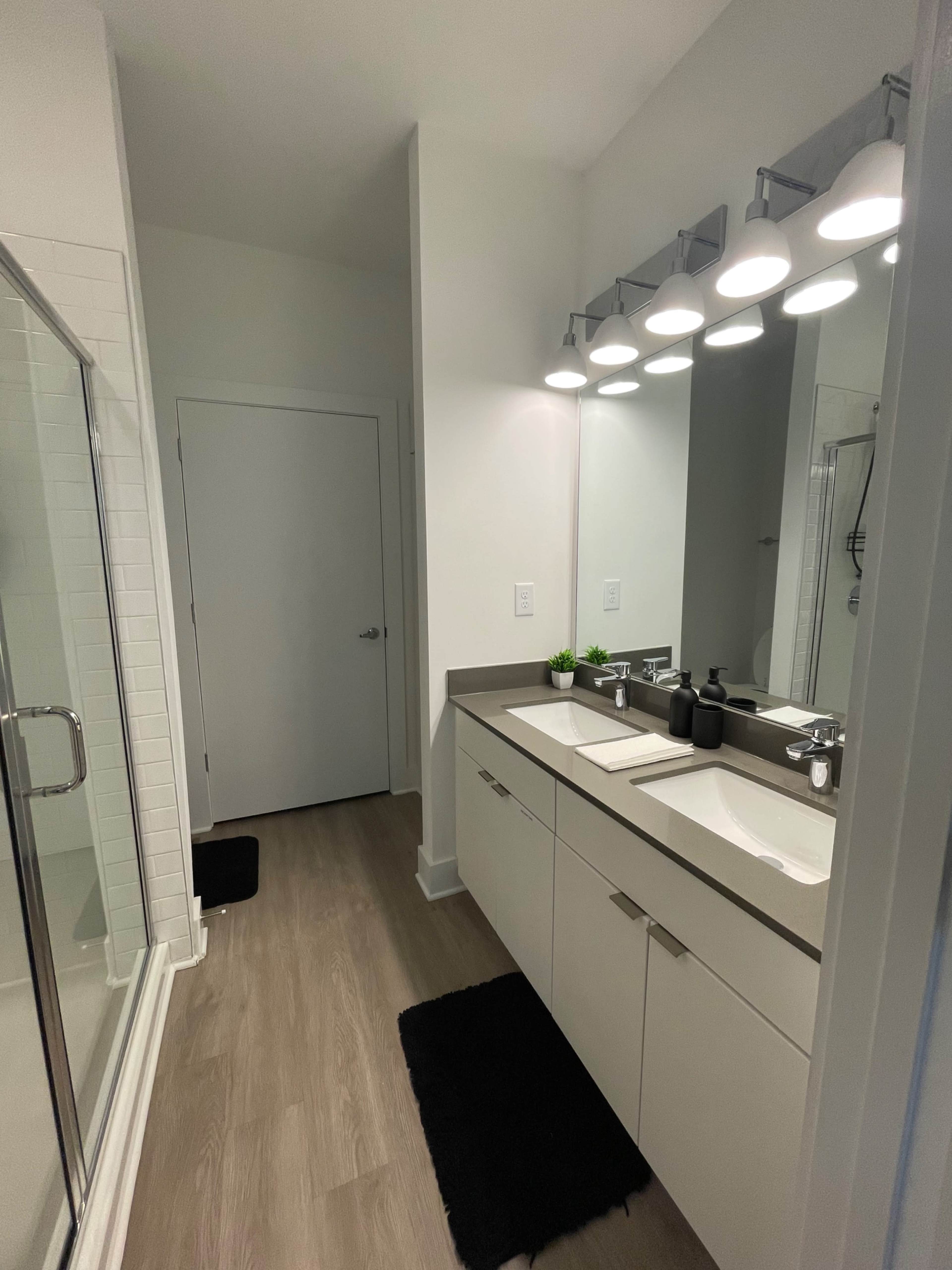 A modern bathroom features a double vanity with two sinks, a large mirror, and a glass shower stall, alongside a closed door and minimal decor.
