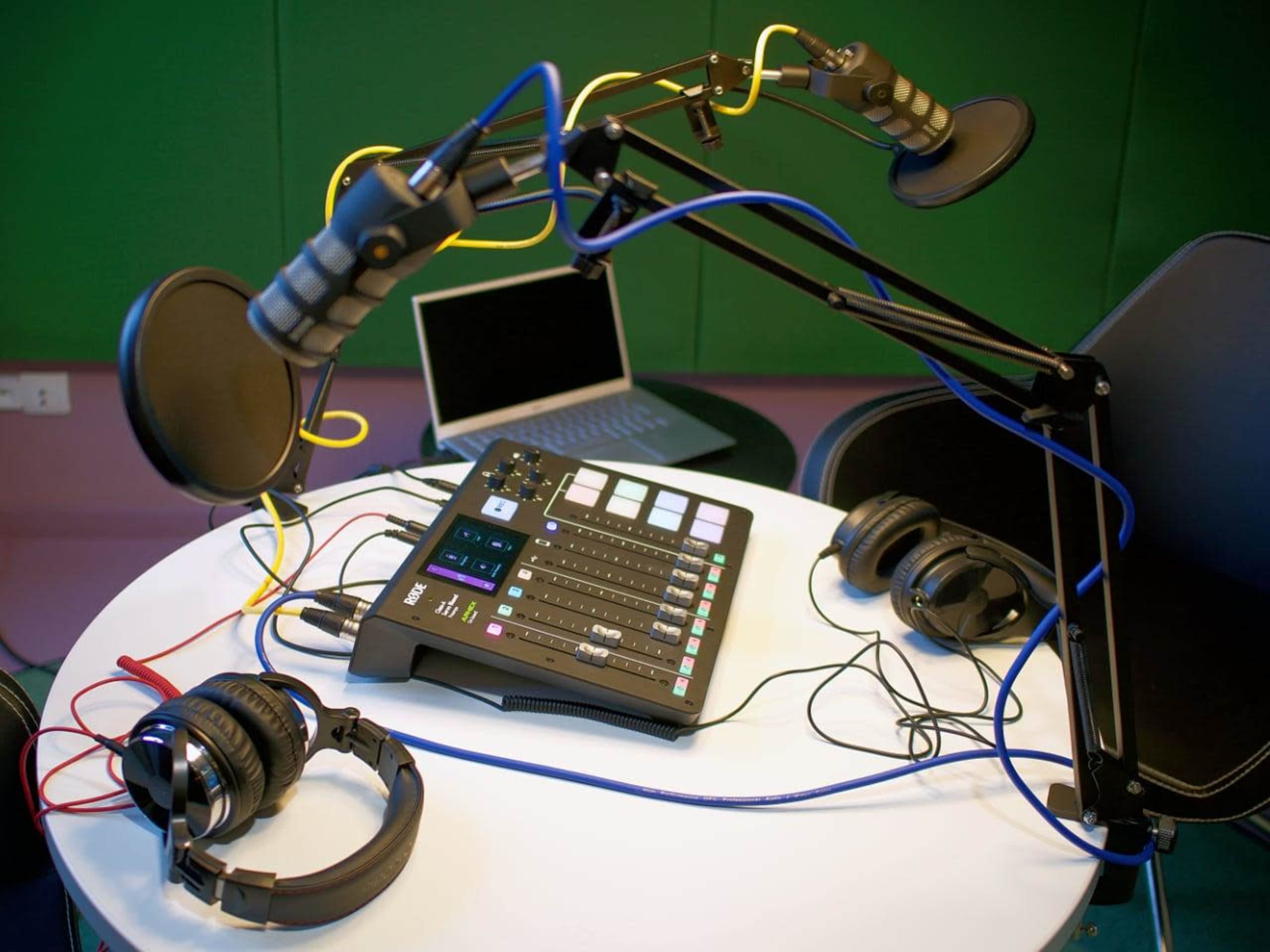The image shows a recording setup featuring two microphone arms, a sound mixer with various controls, and three sets of headphones arranged on a table.