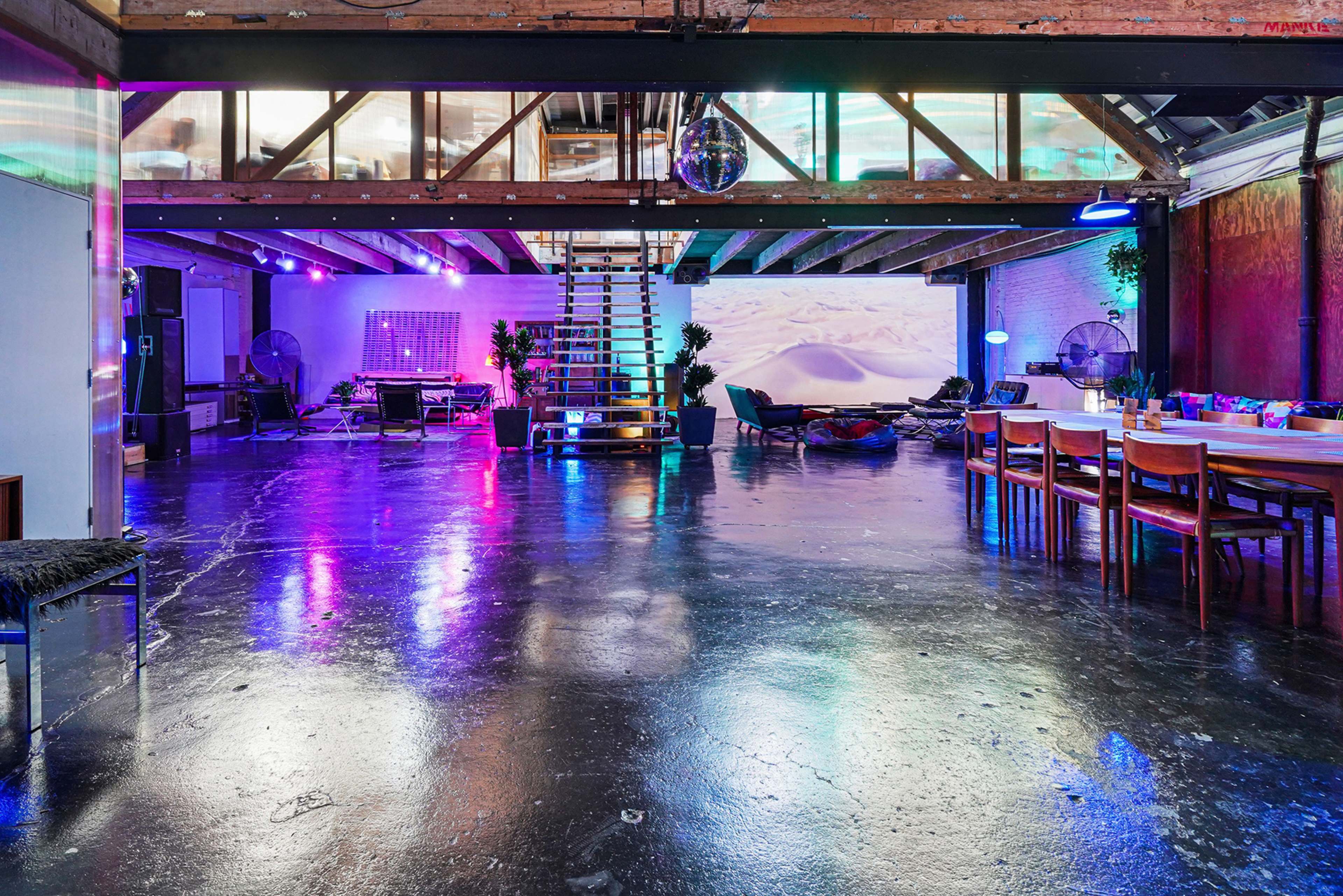 A spacious, modern interior with colorful ambient lighting, exposed beams, and a mix of seating areas featuring a dining table, lounge chairs, and a staircase.