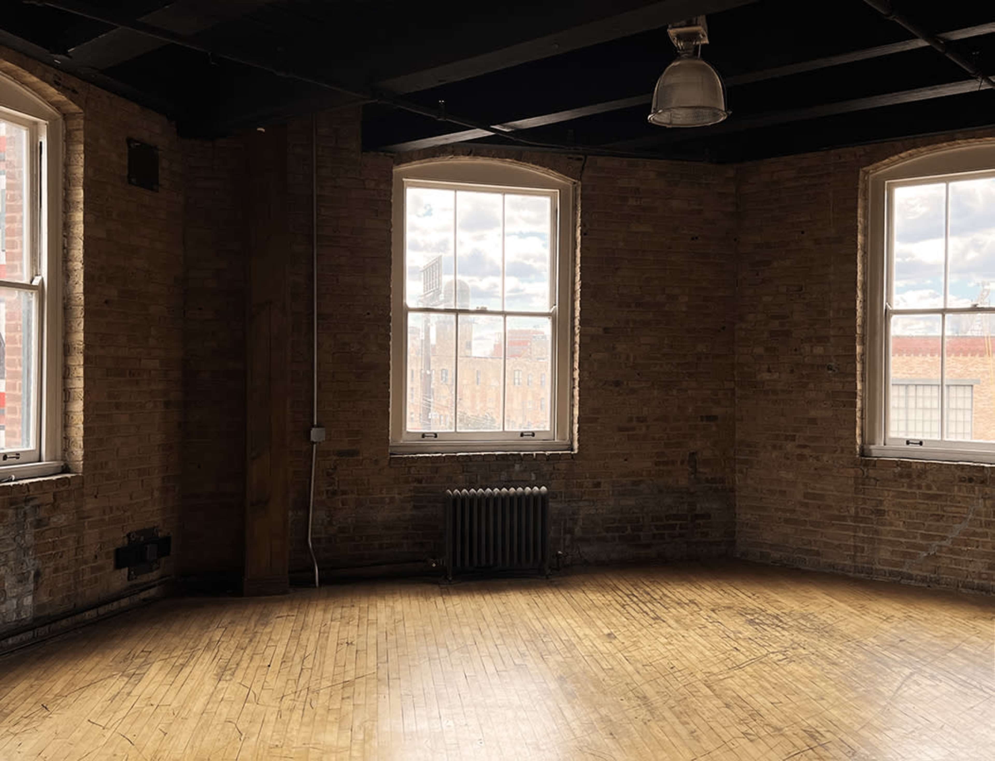 An empty, industrial room with large windows and exposed brick walls, featuring wooden flooring and a radiator.