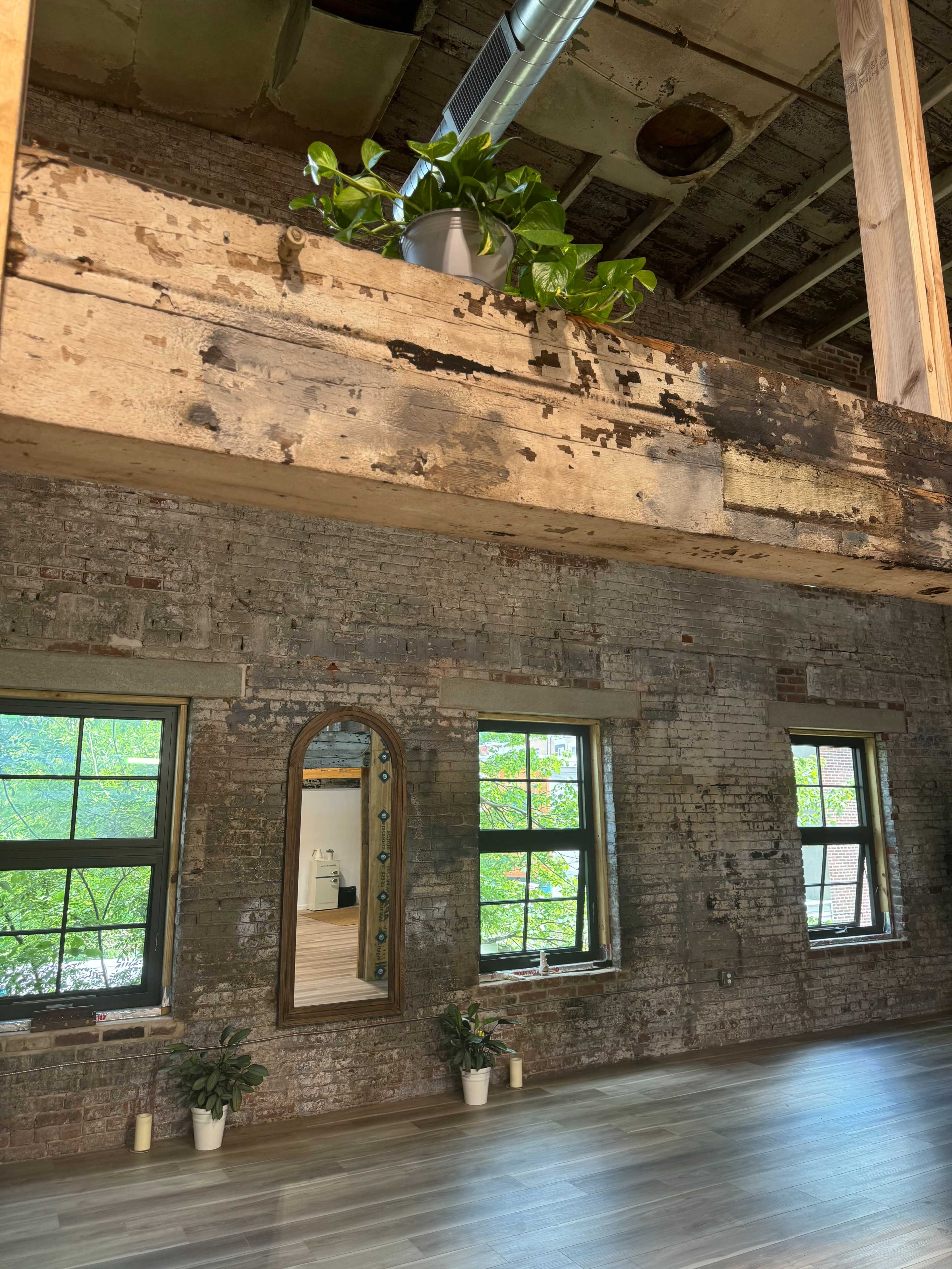 A rustic interior with exposed brick walls, wooden beams, large windows, and a potted plant on a ledge.