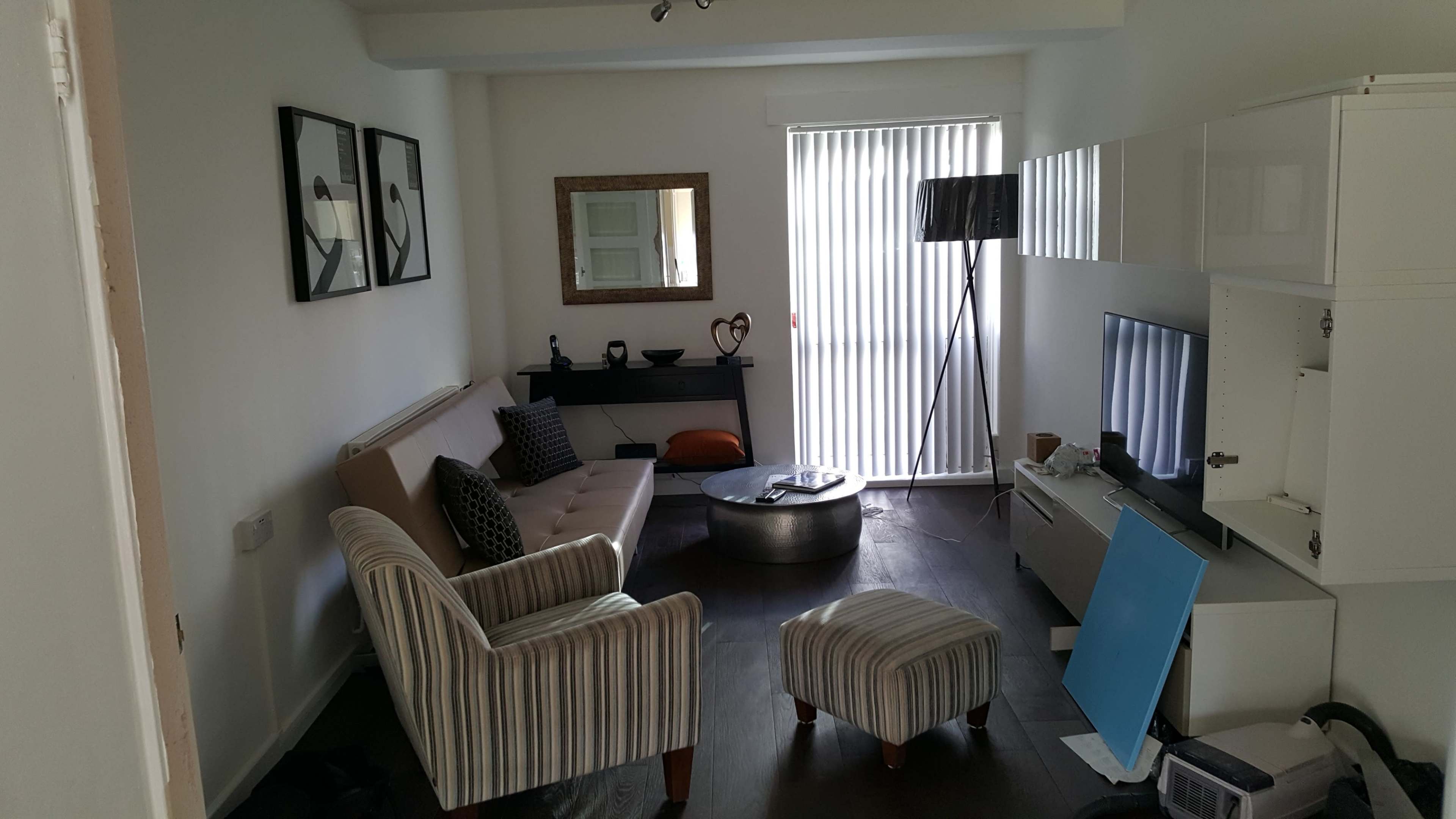 A living room features a beige sofa, a striped ottoman, a coffee table, and a television against a white wall with a sliding glass door.