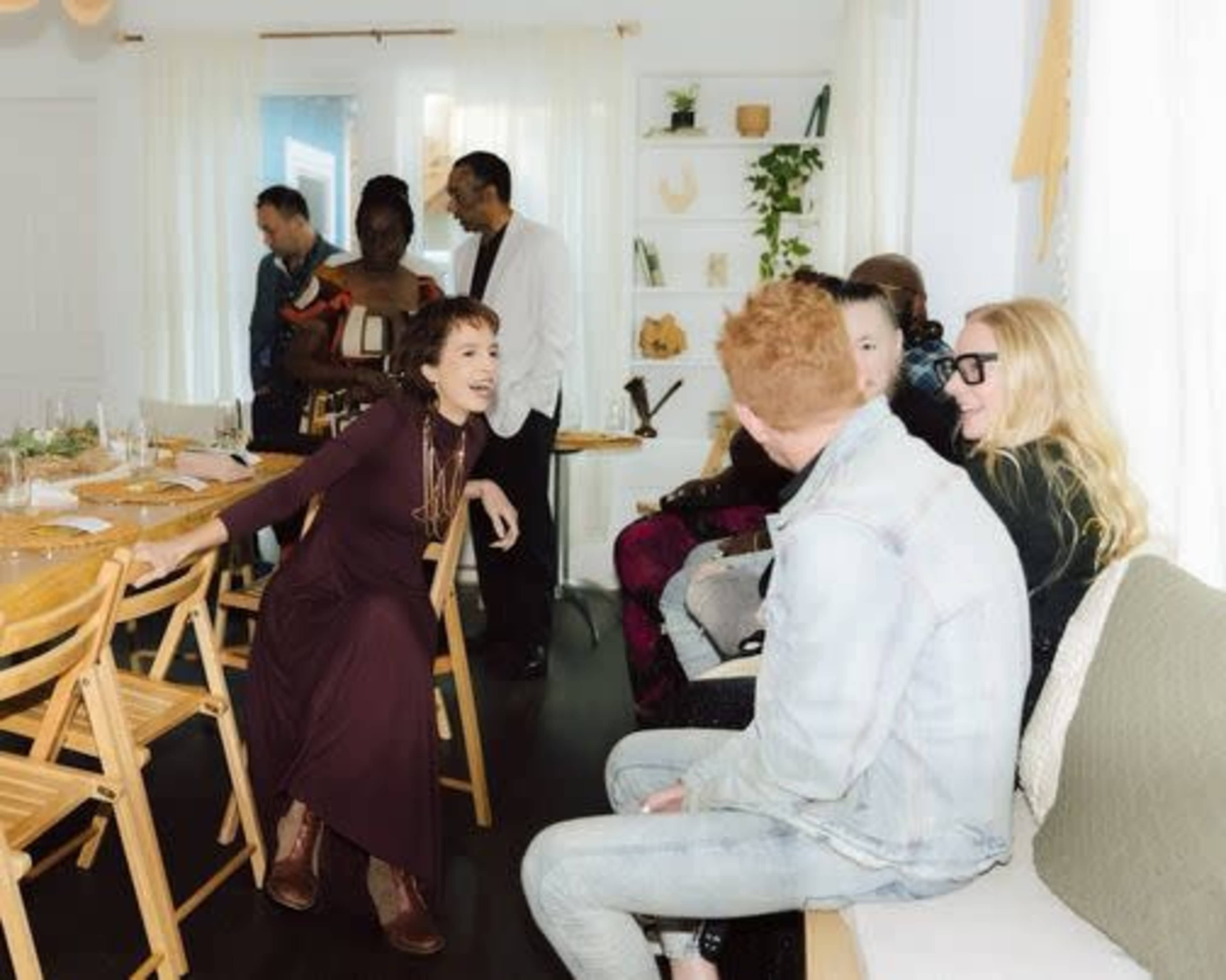 A group of people gathers in a bright room with a dining table, seated and engaging in conversation.