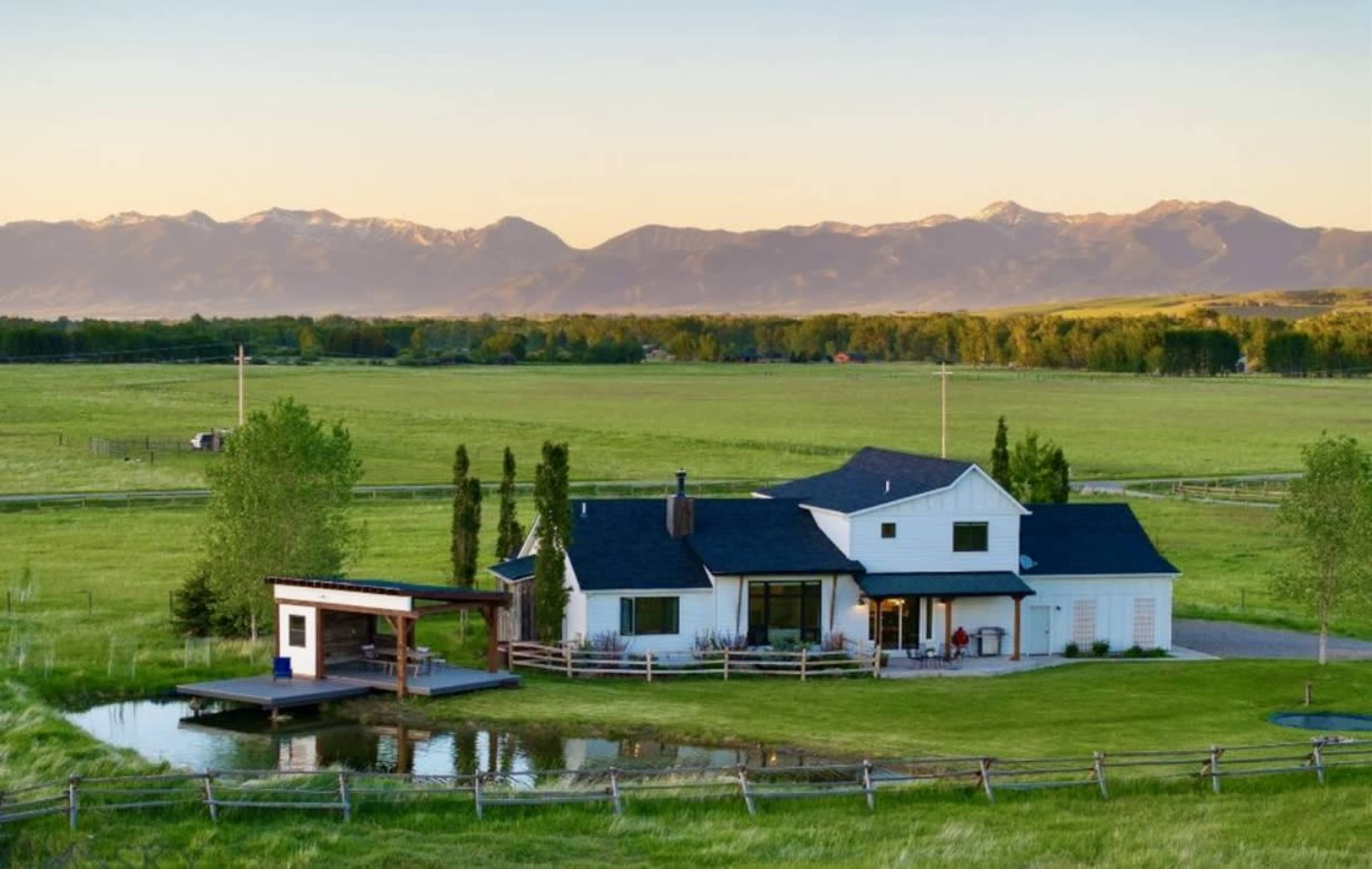 A white house with a black roof sits near a pond, surrounded by green fields and mountains in the background.