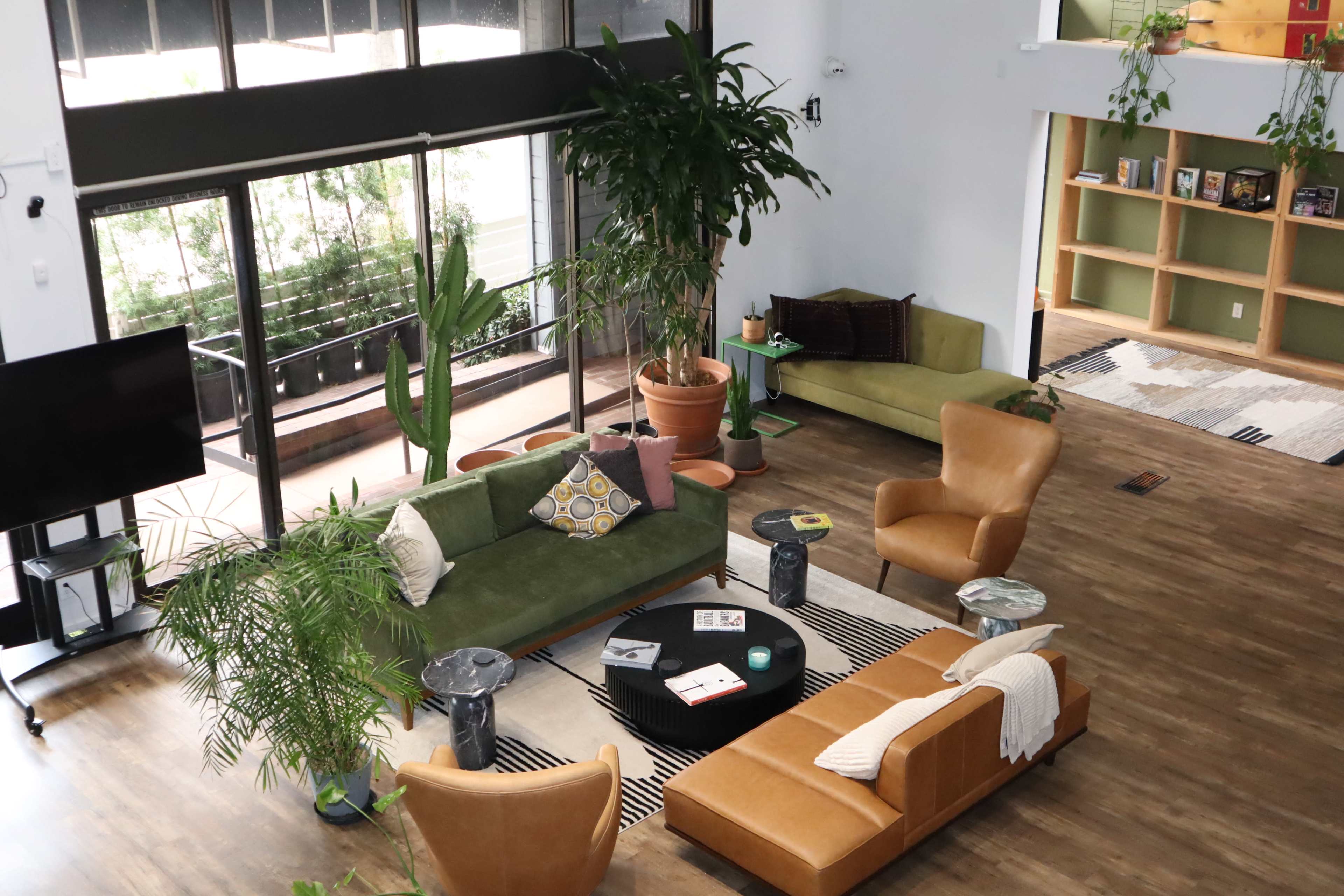 The image shows a spacious living area with large windows, featuring various seating options, potted plants, and a coffee table arranged around a striped rug.