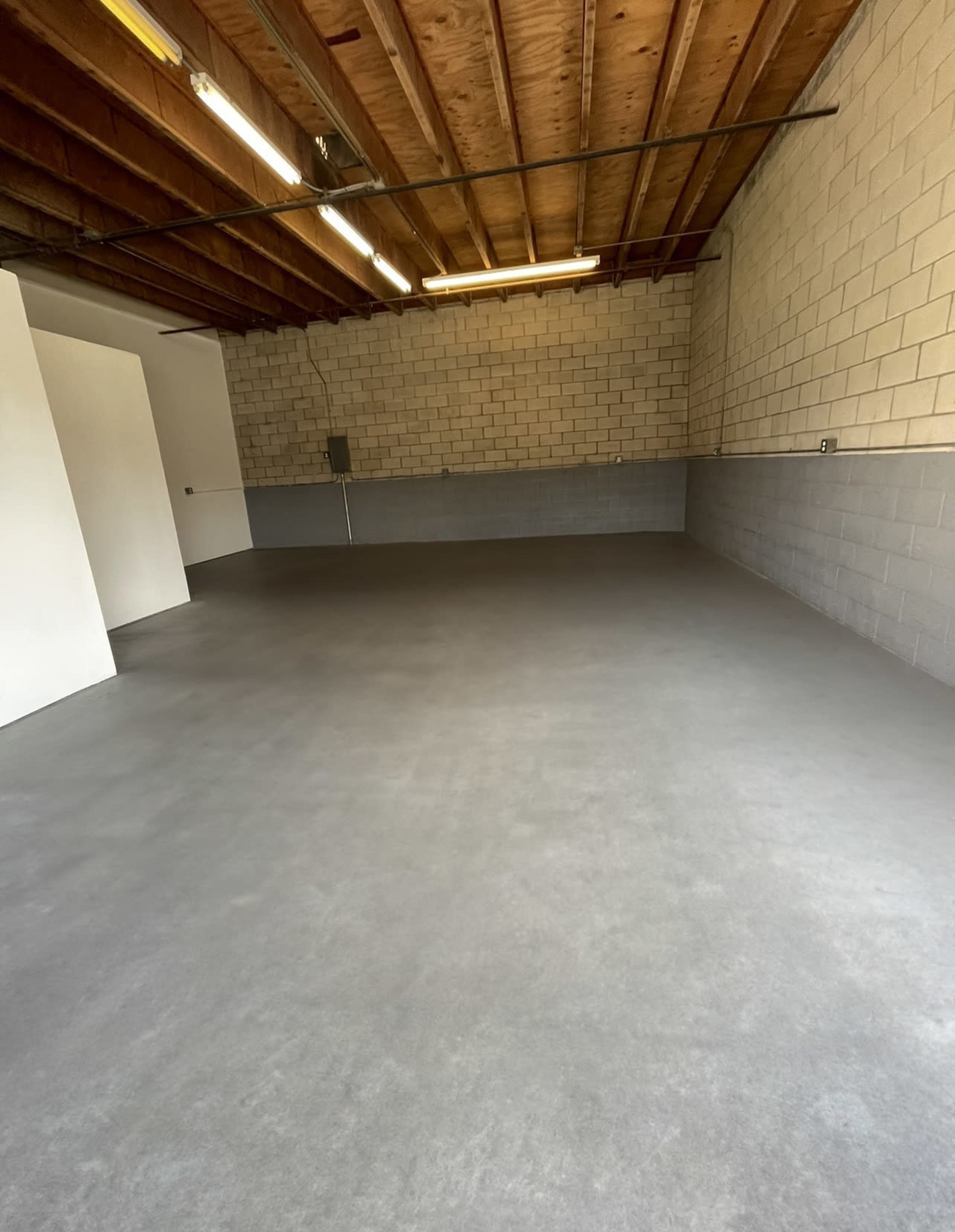 An empty garage or workshop space with concrete flooring, gray cinder block walls, and exposed wooden ceiling beams.
