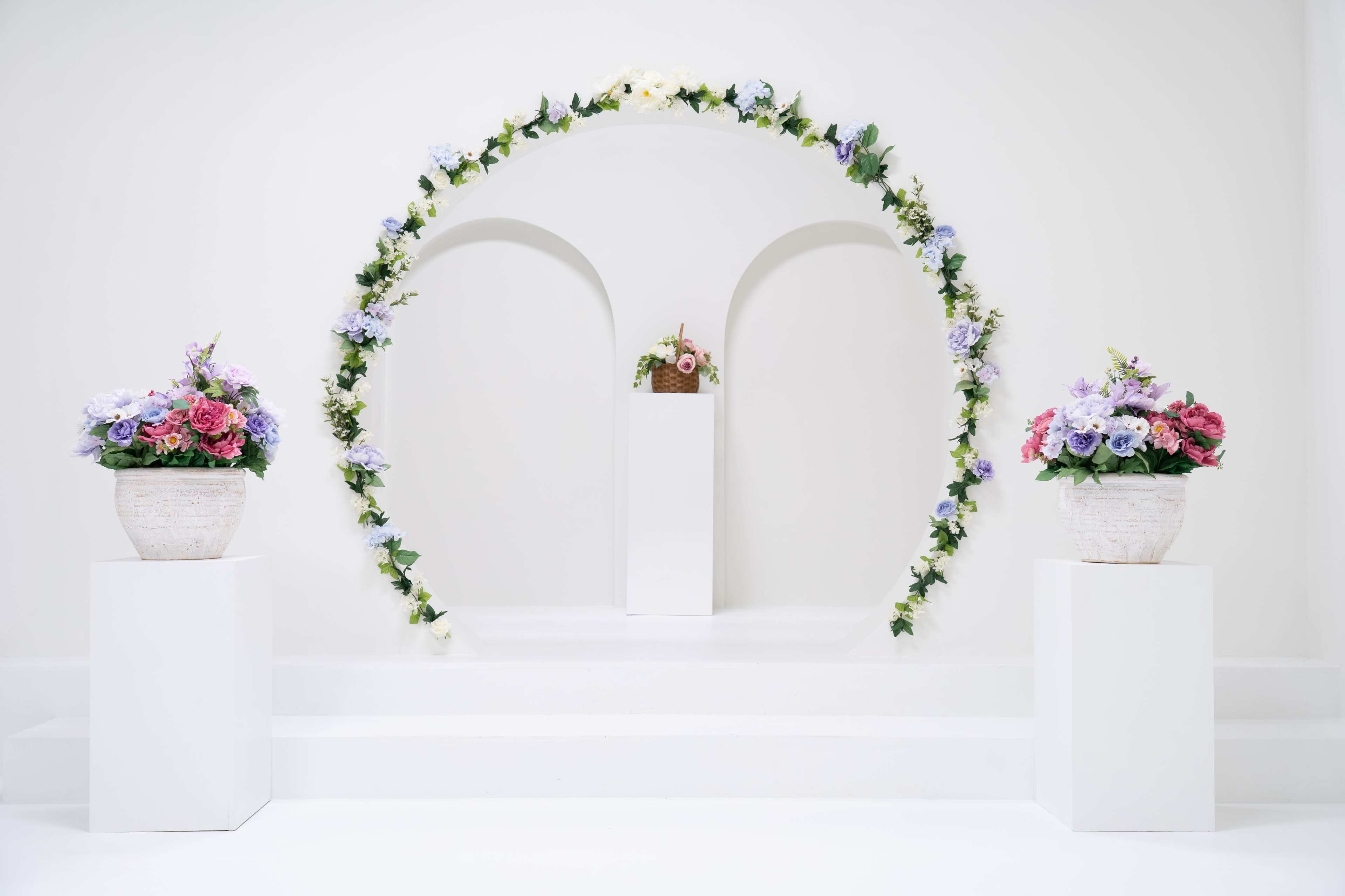 The image features a minimalist wedding altar adorned with a floral arch and two potted flower arrangements on white pedestals.