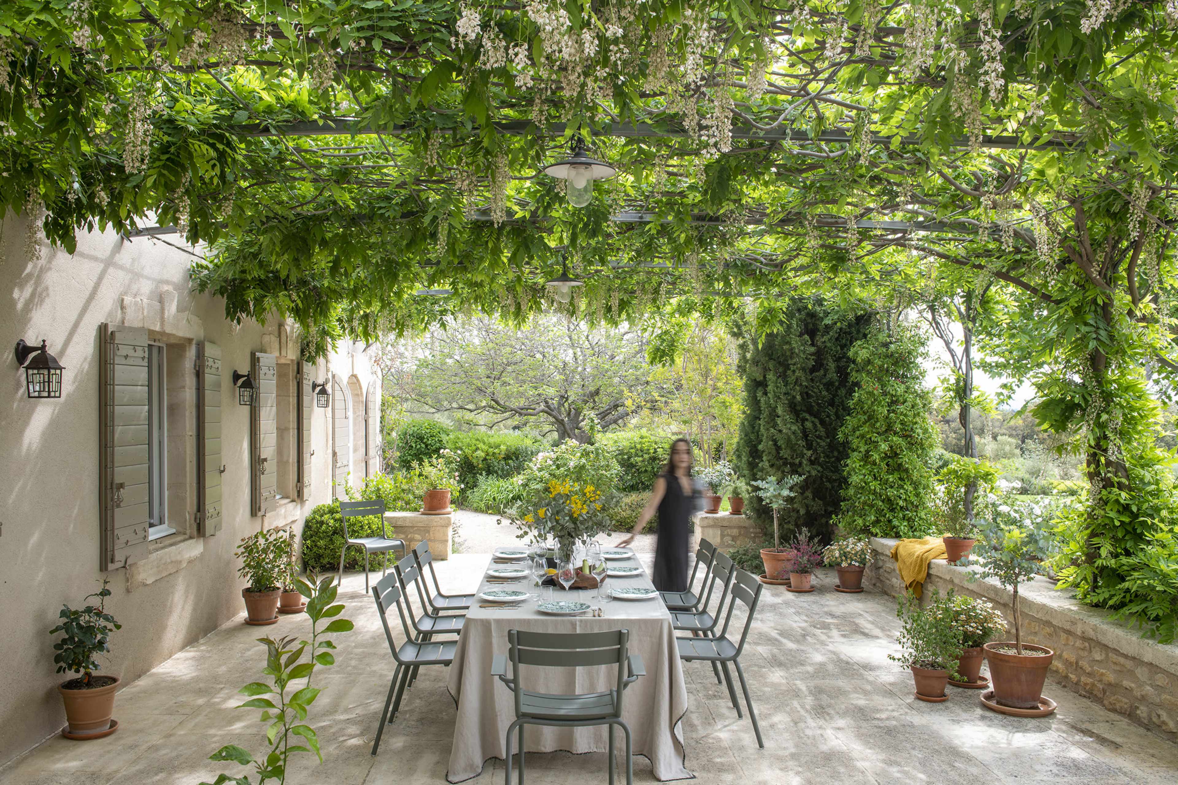 A dining table is set under a pergola covered with lush greenery, surrounded by potted plants and a scenic view in the background.