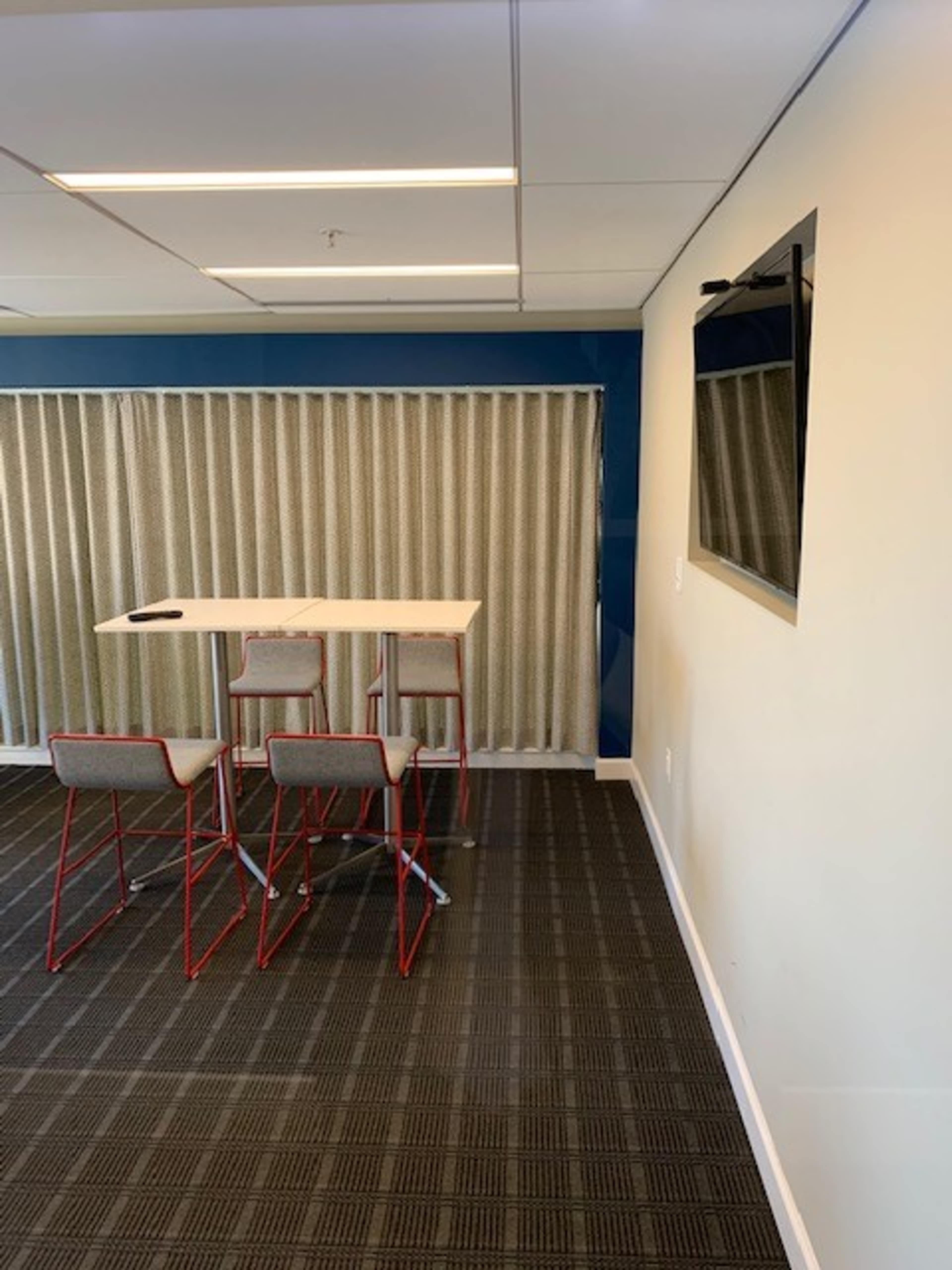 A small meeting area features a high table with four bar stools against a wall with a television and a curtain.