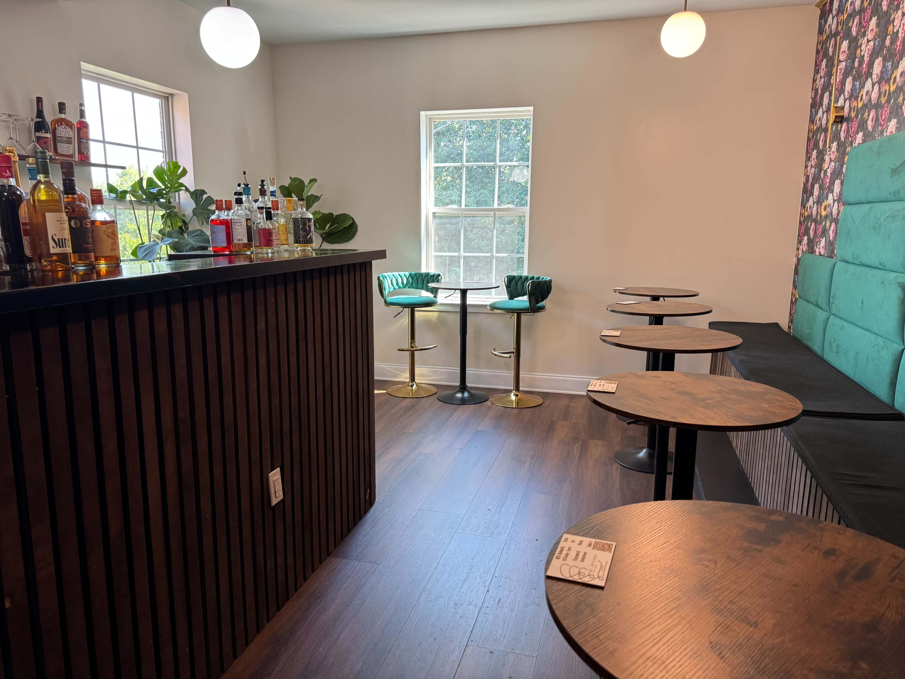 The image shows a modern bar area featuring a black wooden counter, several glass shelves with liquor bottles, and a seating area with green upholstered chairs and round wooden tables.