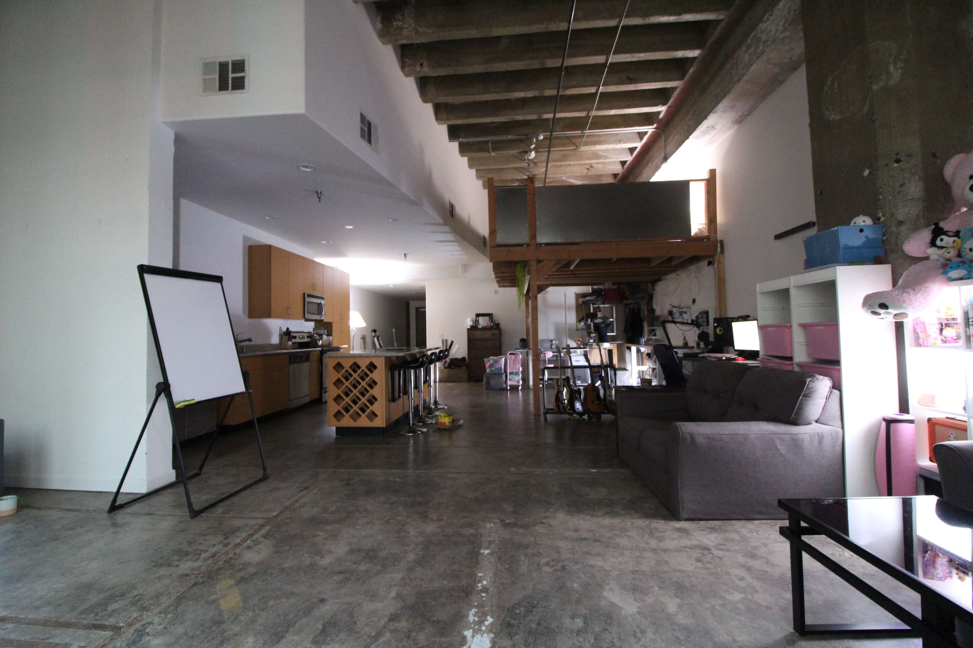 A spacious, industrial-style interior features a kitchen area, a living space with a sofa, and an upper loft space, all illuminated by natural light.