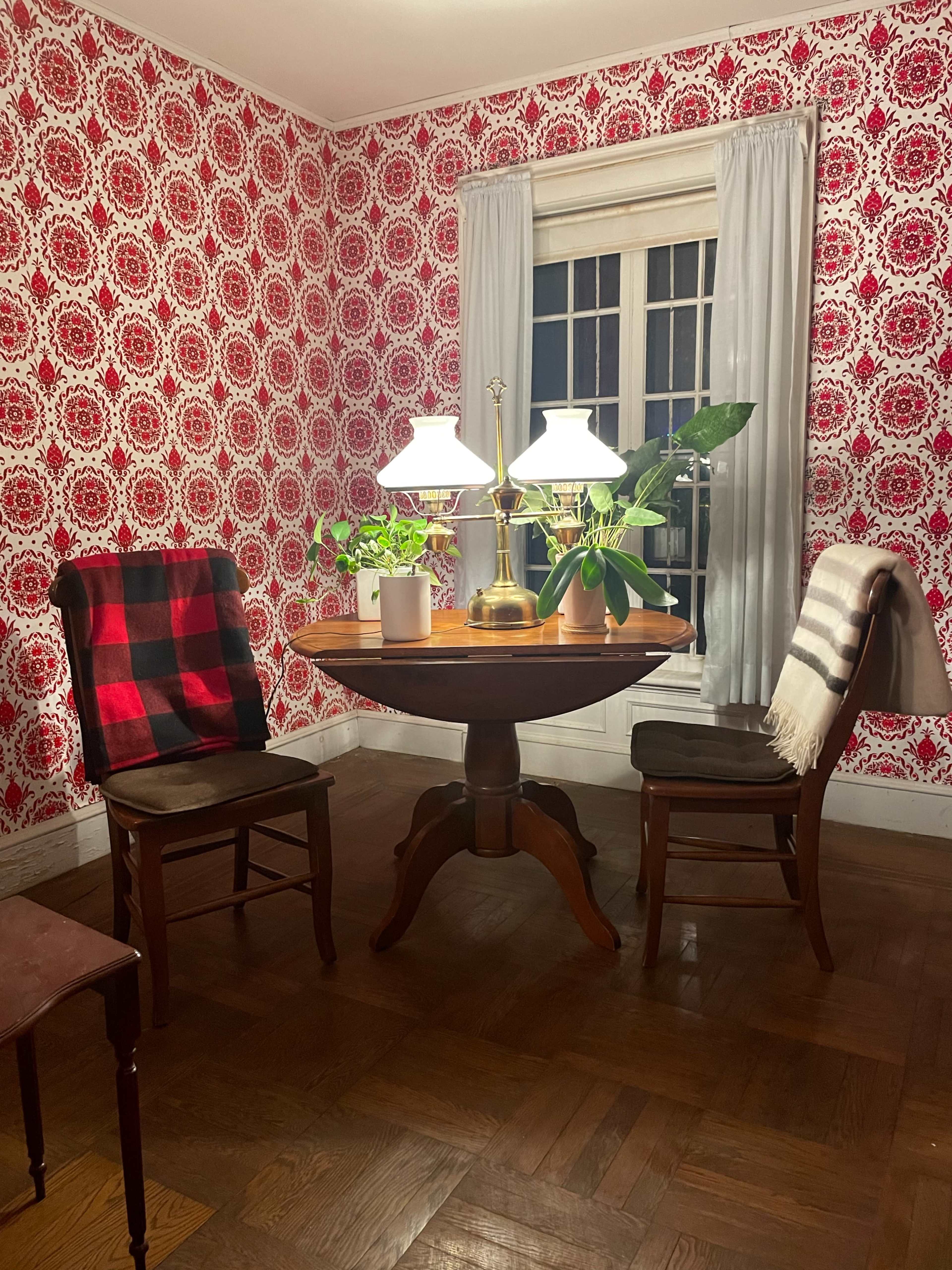 A round wooden table is set between two chairs in a room with floral wallpaper, illuminated by two lamps and accented with potted plants.