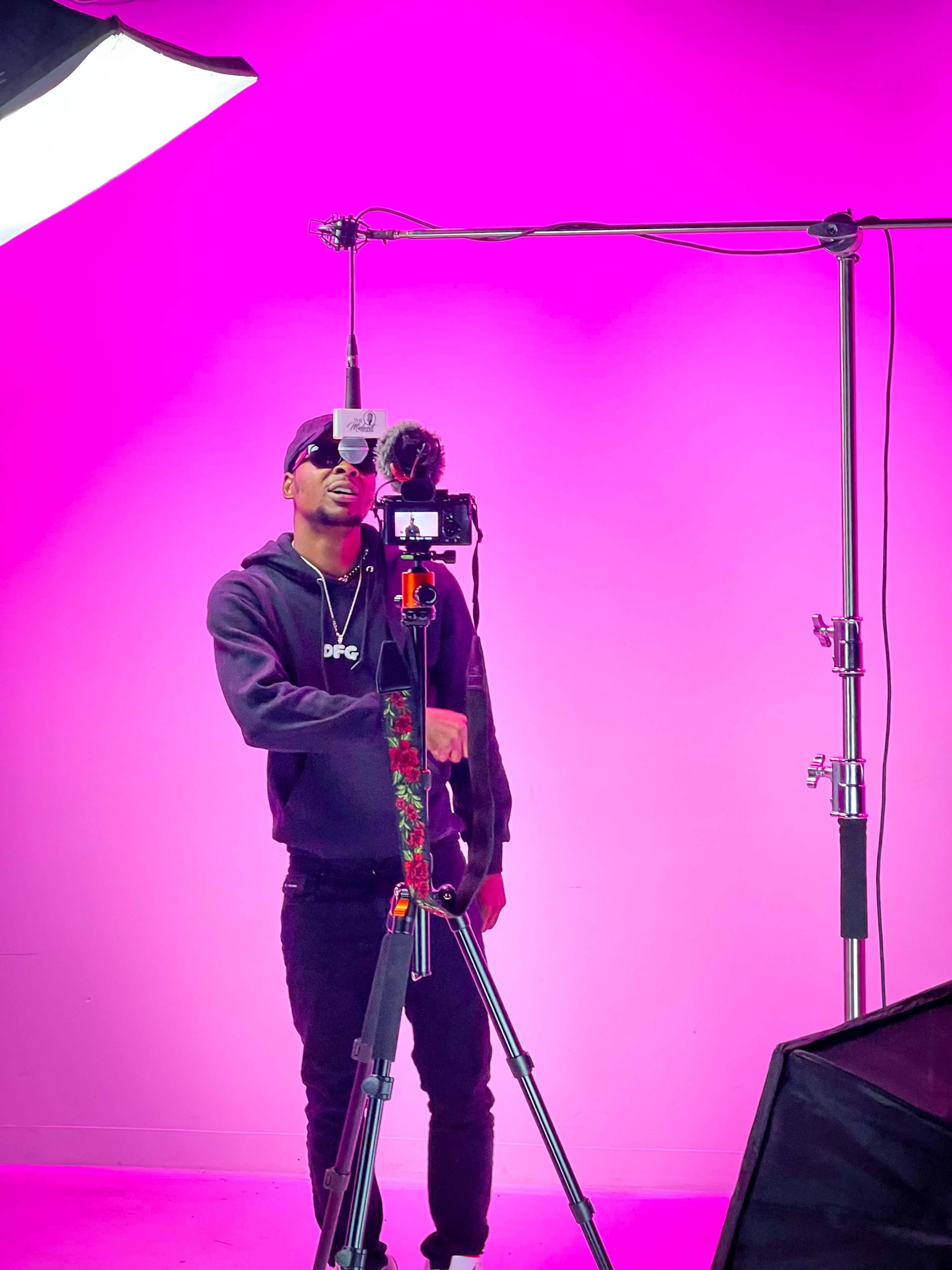 A person stands in front of a bright pink backdrop, adjusting a camera mounted on a tripod with a microphone attached.