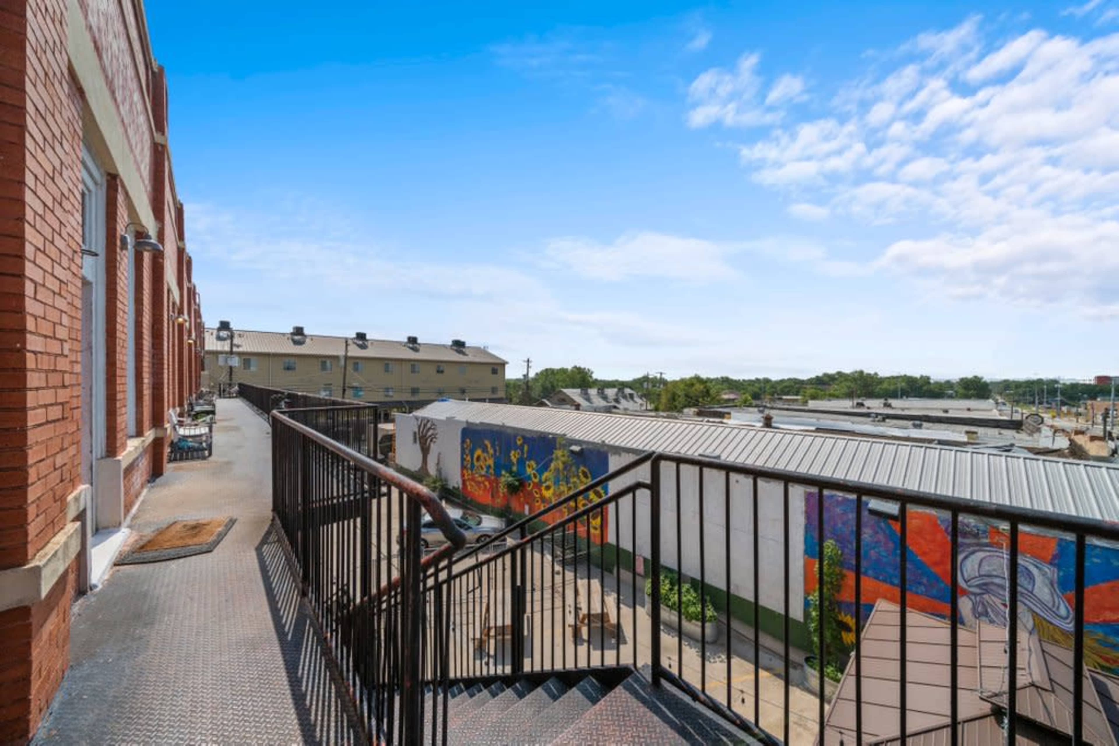 The image shows a metal staircase leading down from a second-story balcony, with a view of nearby buildings and a colorful mural on a wall.