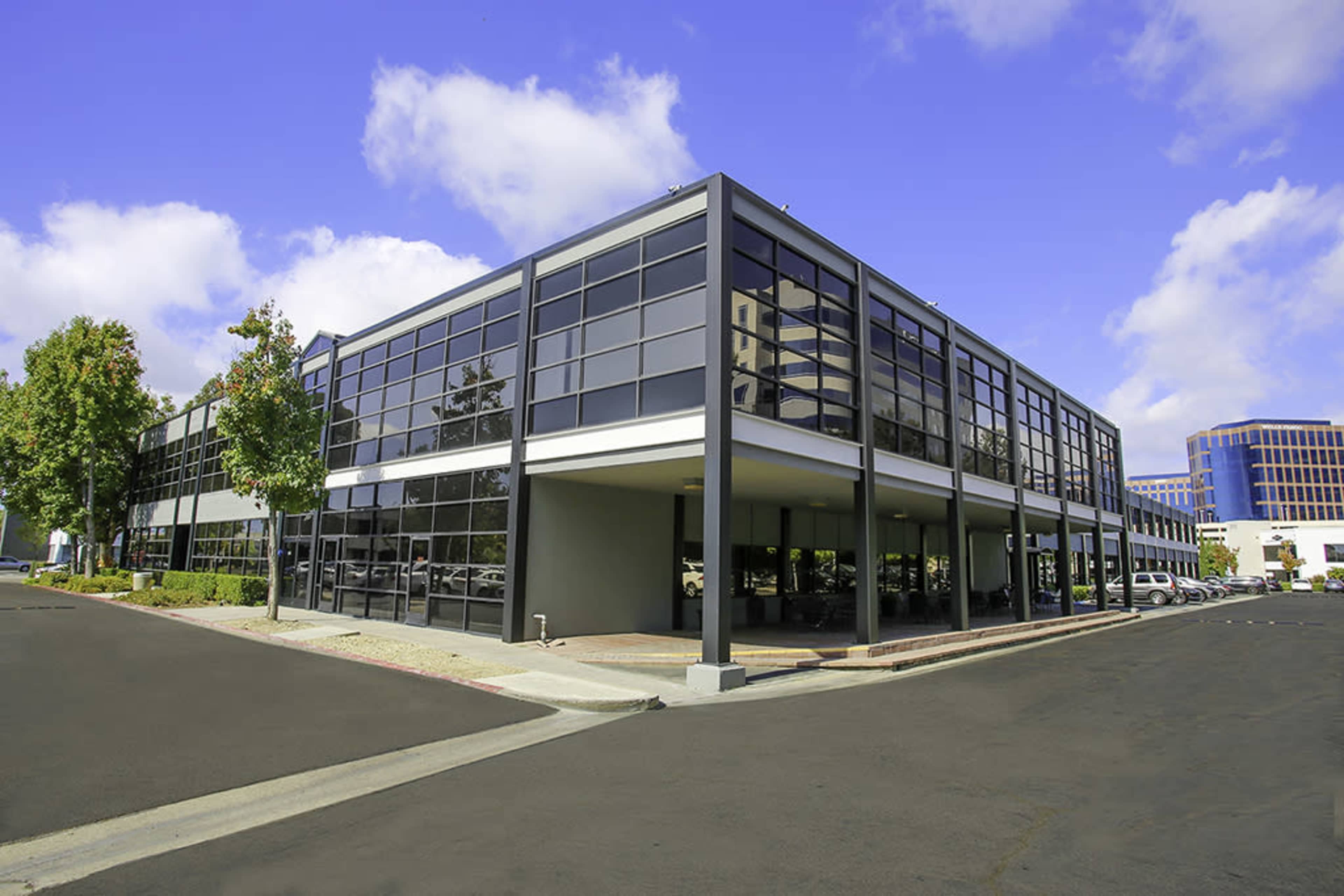 A modern office building with large glass windows is situated on a paved lot under a clear blue sky.