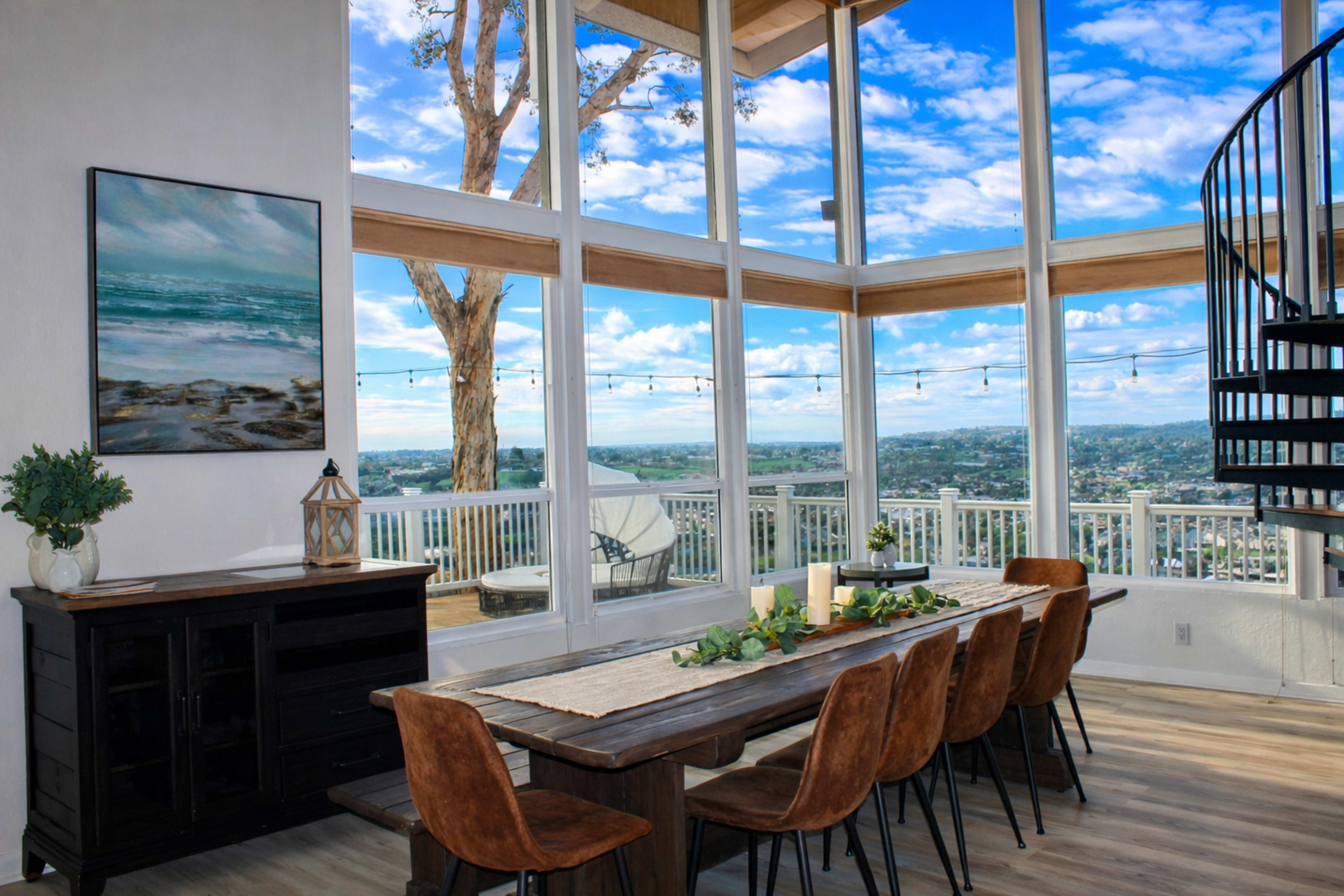 A spacious dining area with a large wooden table, surrounded by chairs, features large windows that offer a view of the sky and landscape outside.