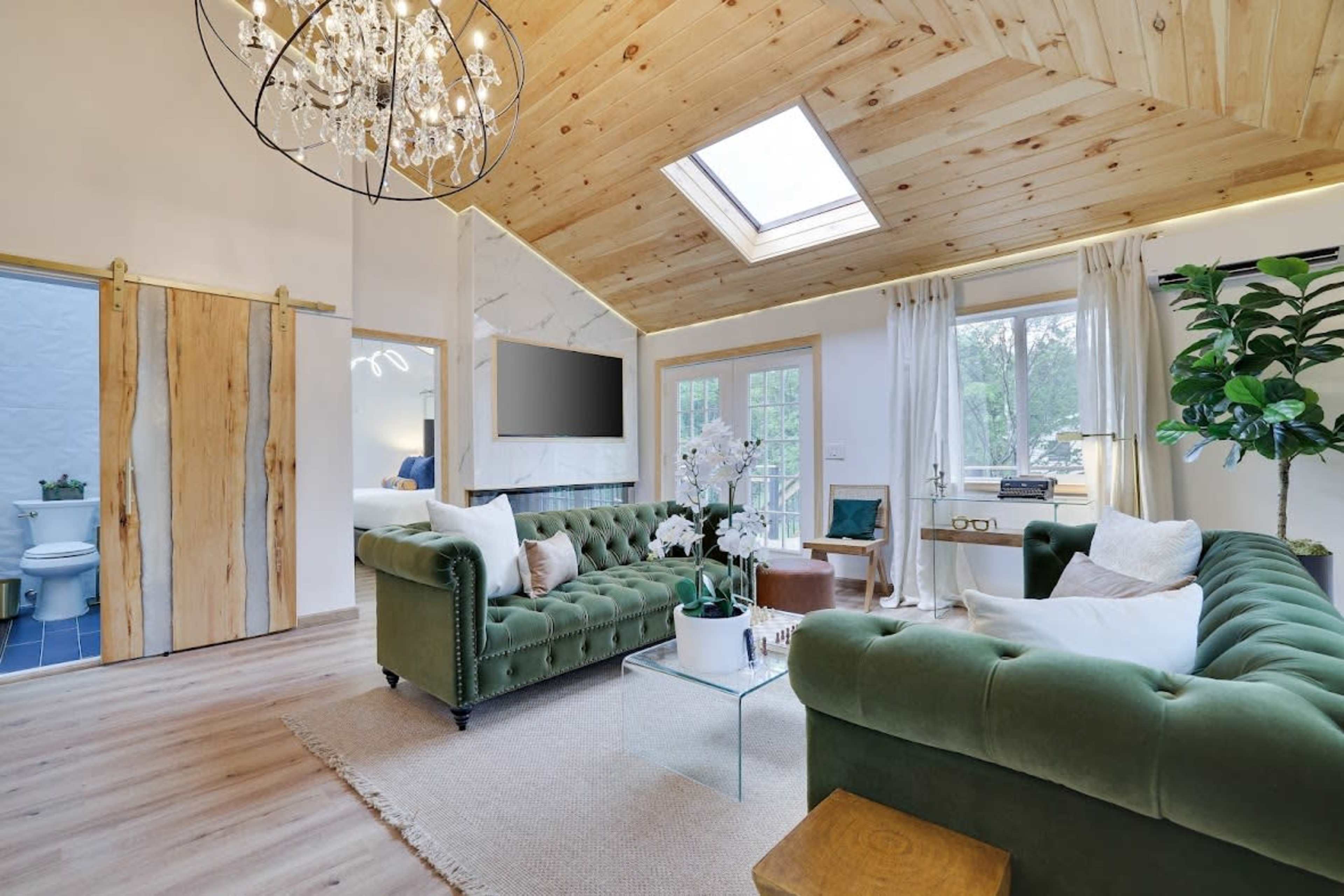 The image shows a stylish living room with a green upholstered sofa, a glass coffee table, and a large skylight under a wooden ceiling.