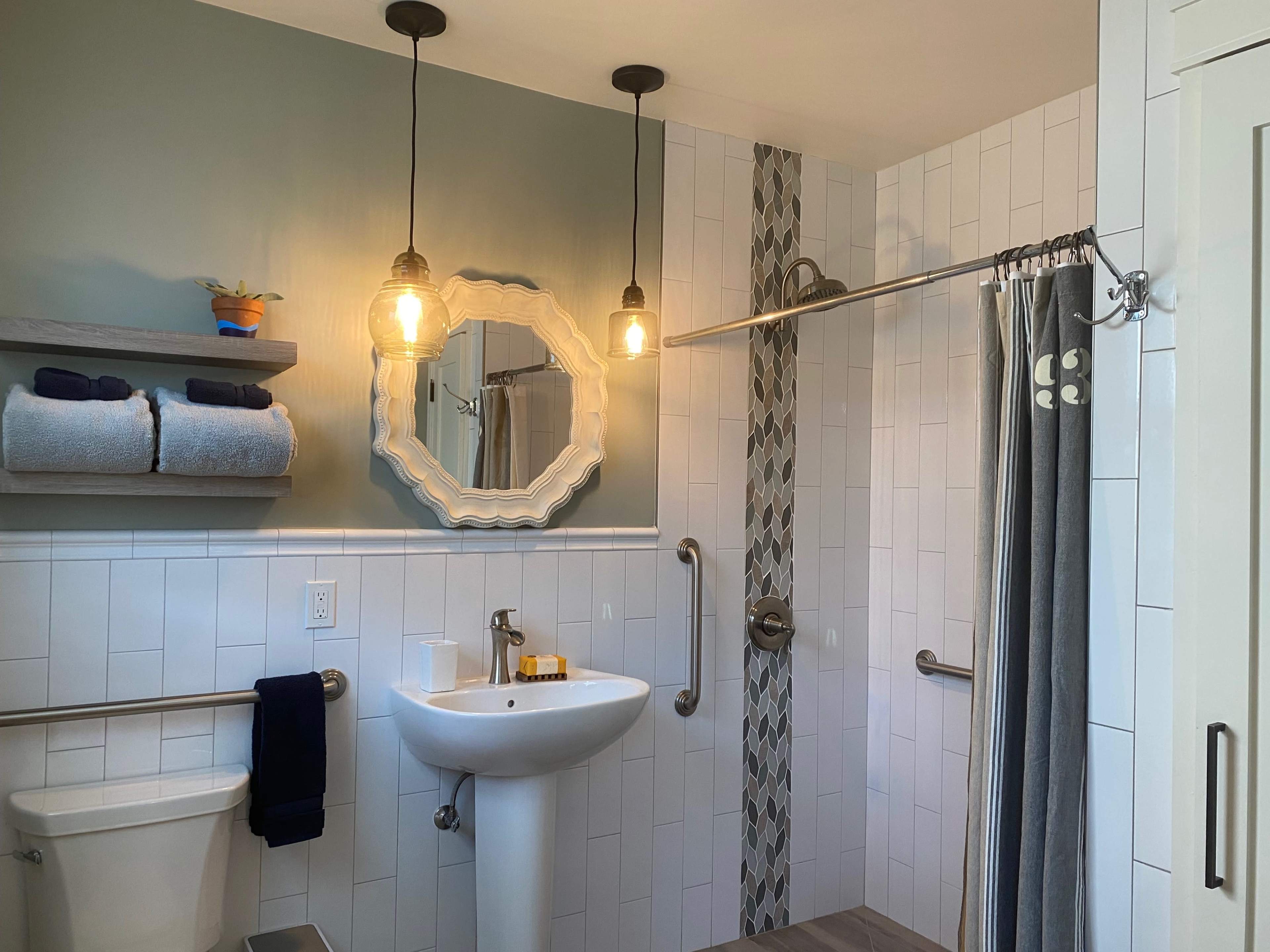 The image shows a modern bathroom featuring a walk-in shower with a wall-mounted grab bar, a pedestal sink, a large mirror, and a shelf holding neatly folded towels.