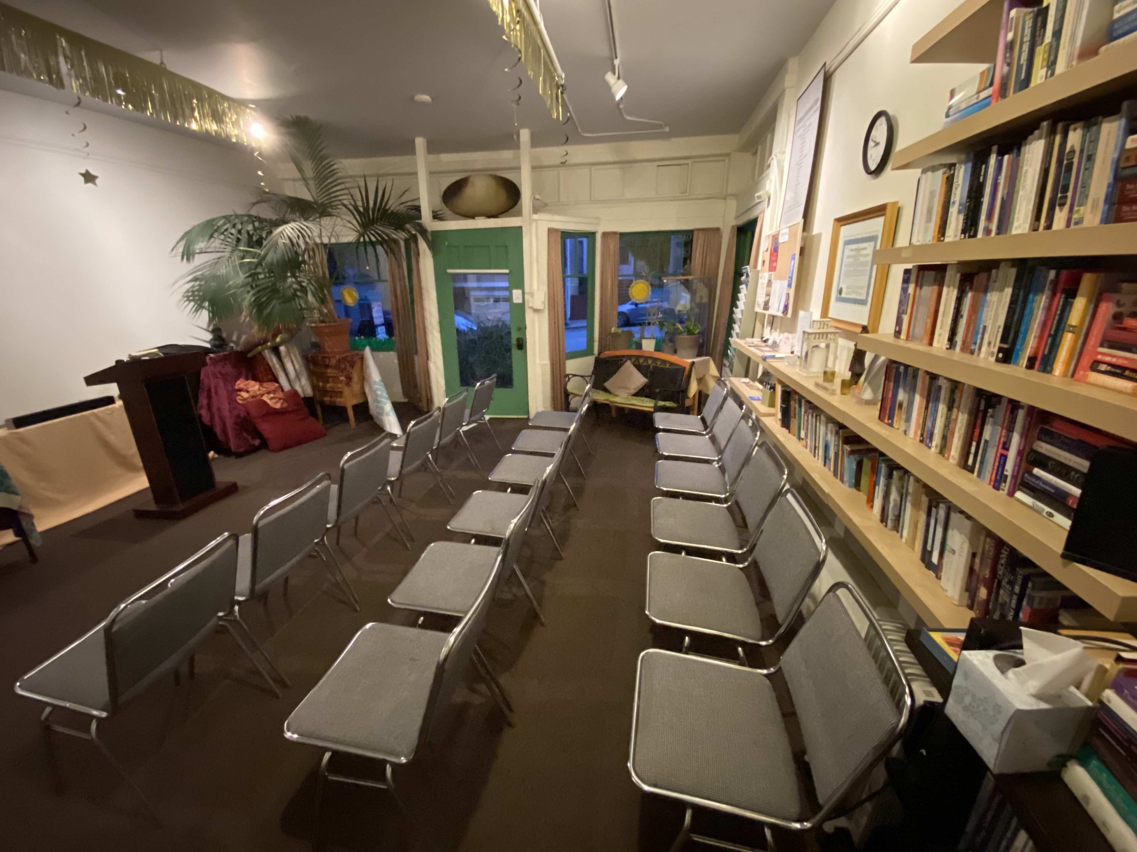 A room with several rows of metal chairs facing a small podium, alongside bookshelves filled with books and a large window letting in natural light.