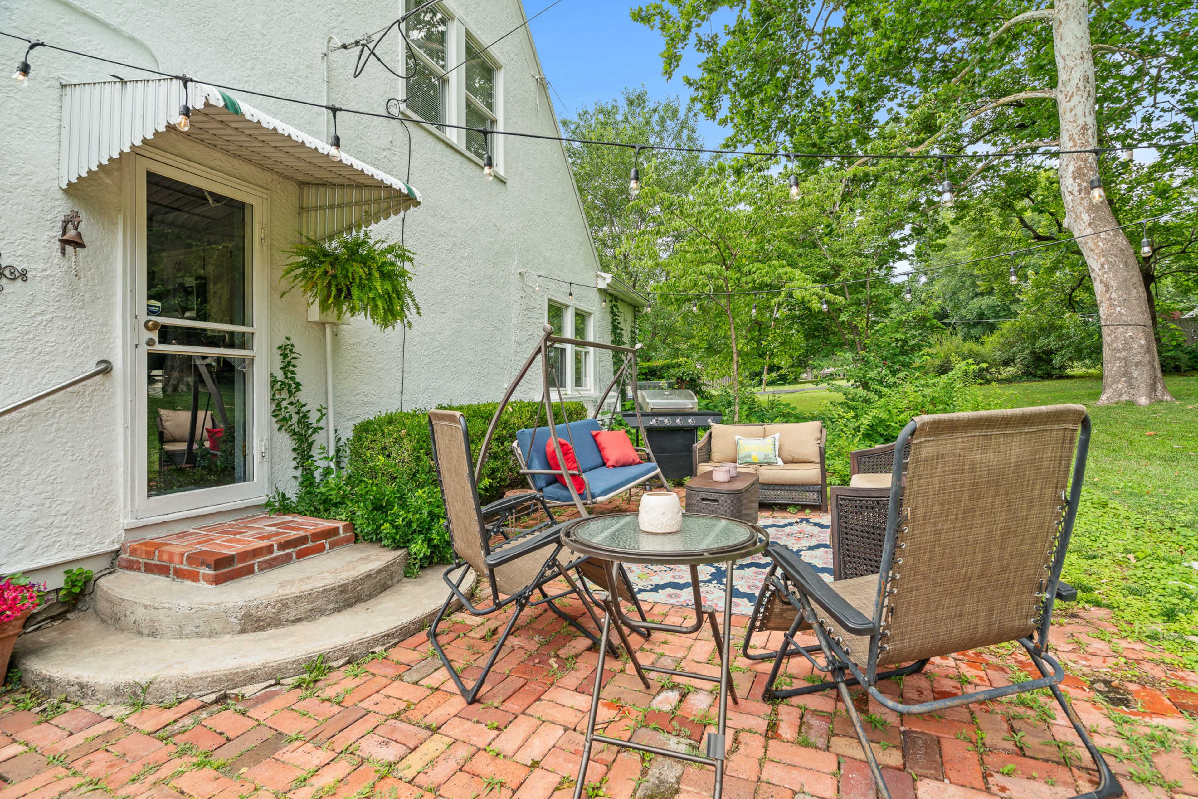 A cozy outdoor seating area features a table and chairs on a brick patio surrounded by greenery and adorned with decorative string lights.