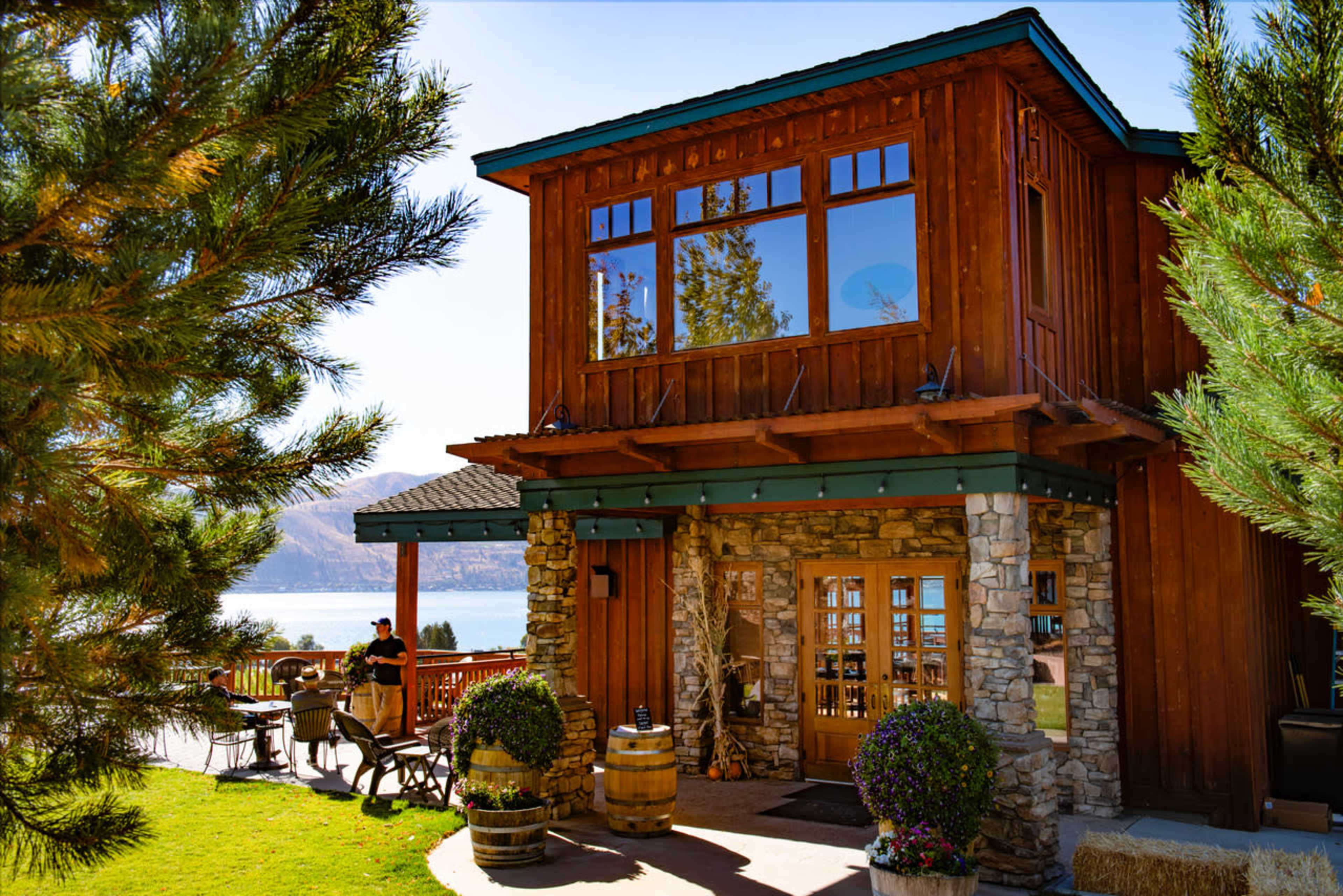 A two-story wooden building with large windows sits near a lake, surrounded by trees and outdoor seating.