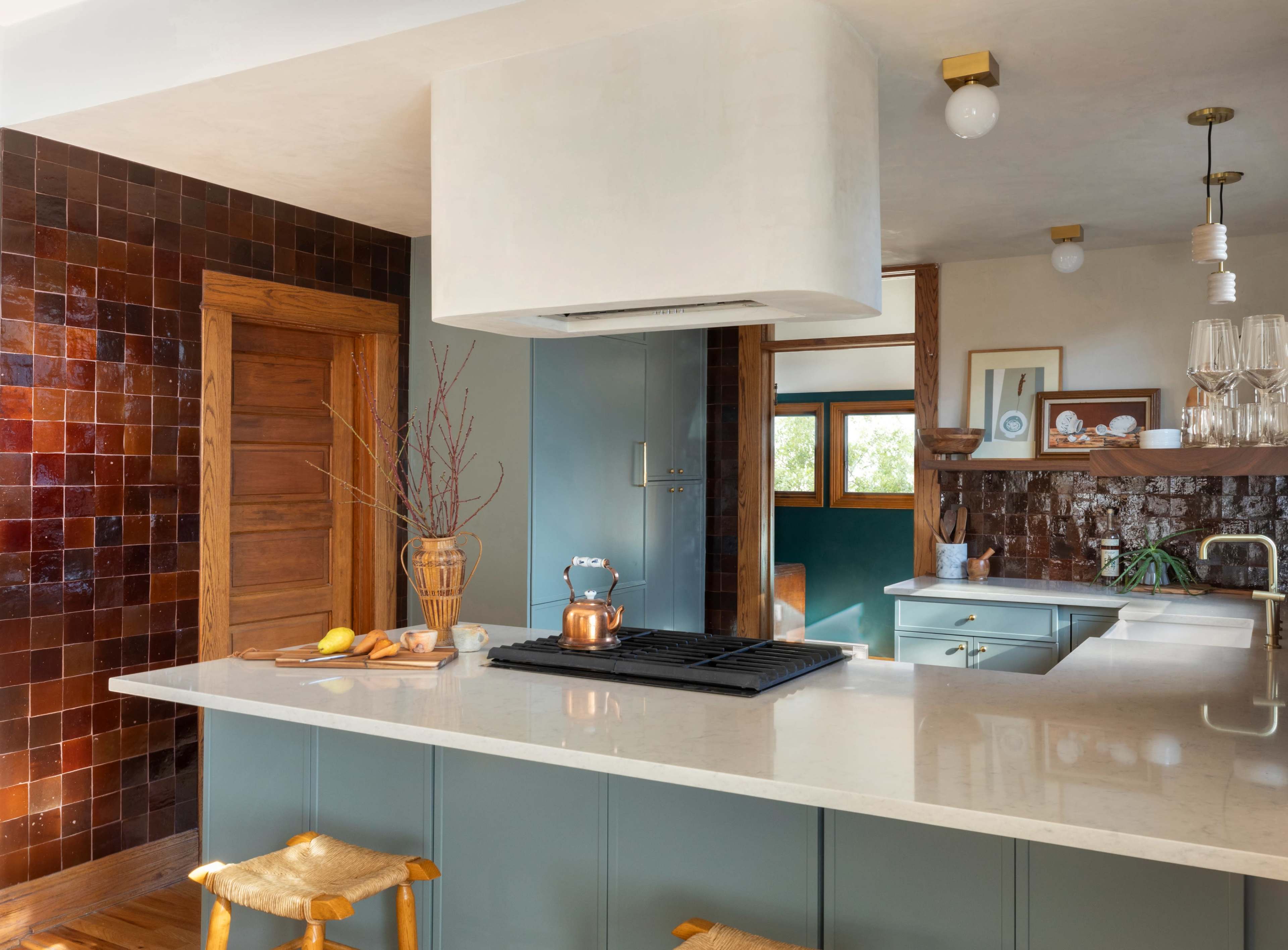 A modern kitchen features a central island with a cooktop, surrounded by textured walls of dark tiles and wooden accents.