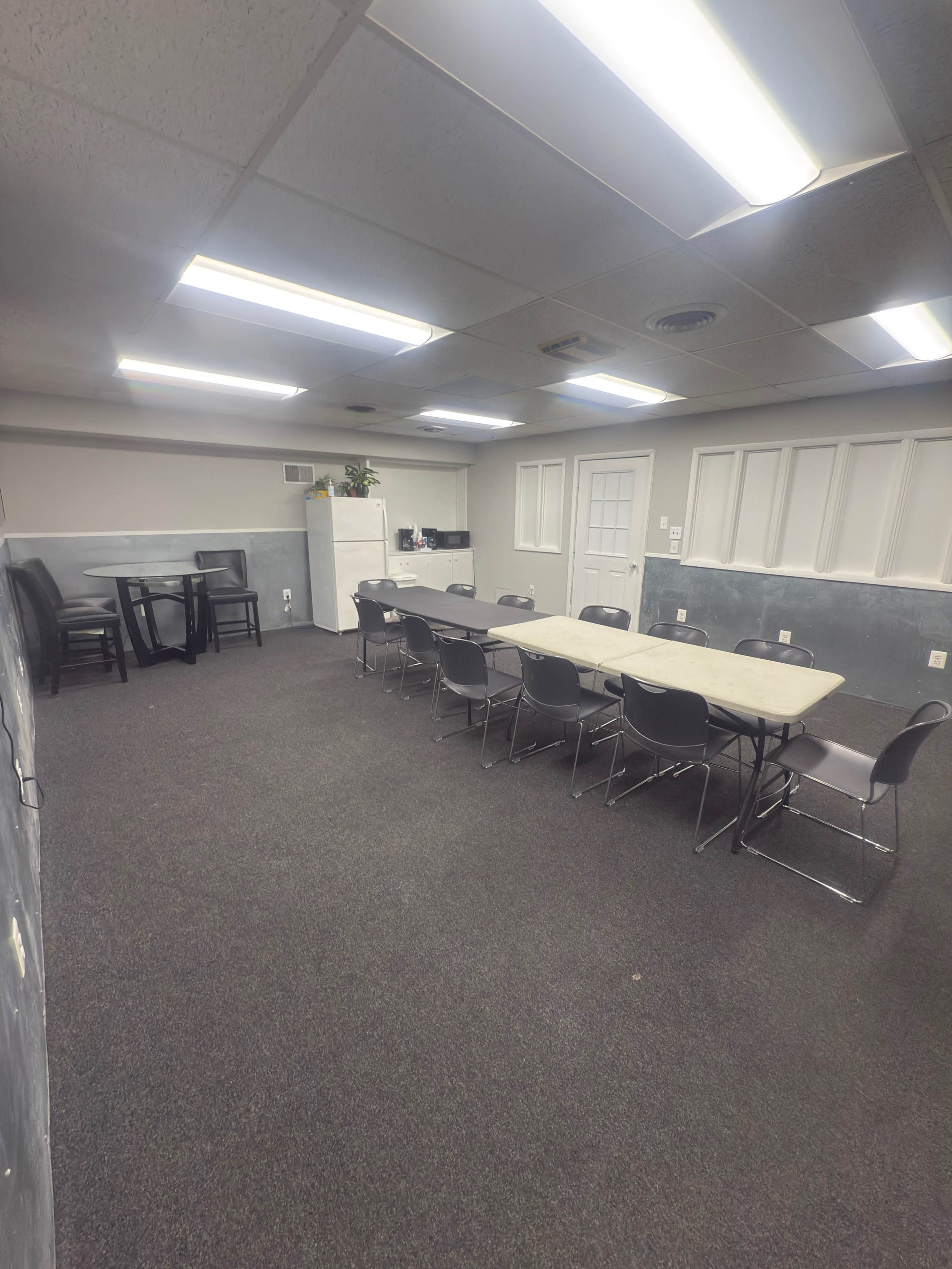The image shows a spacious meeting room with a large table surrounded by chairs, a kitchenette area in the background, and gray walls.
