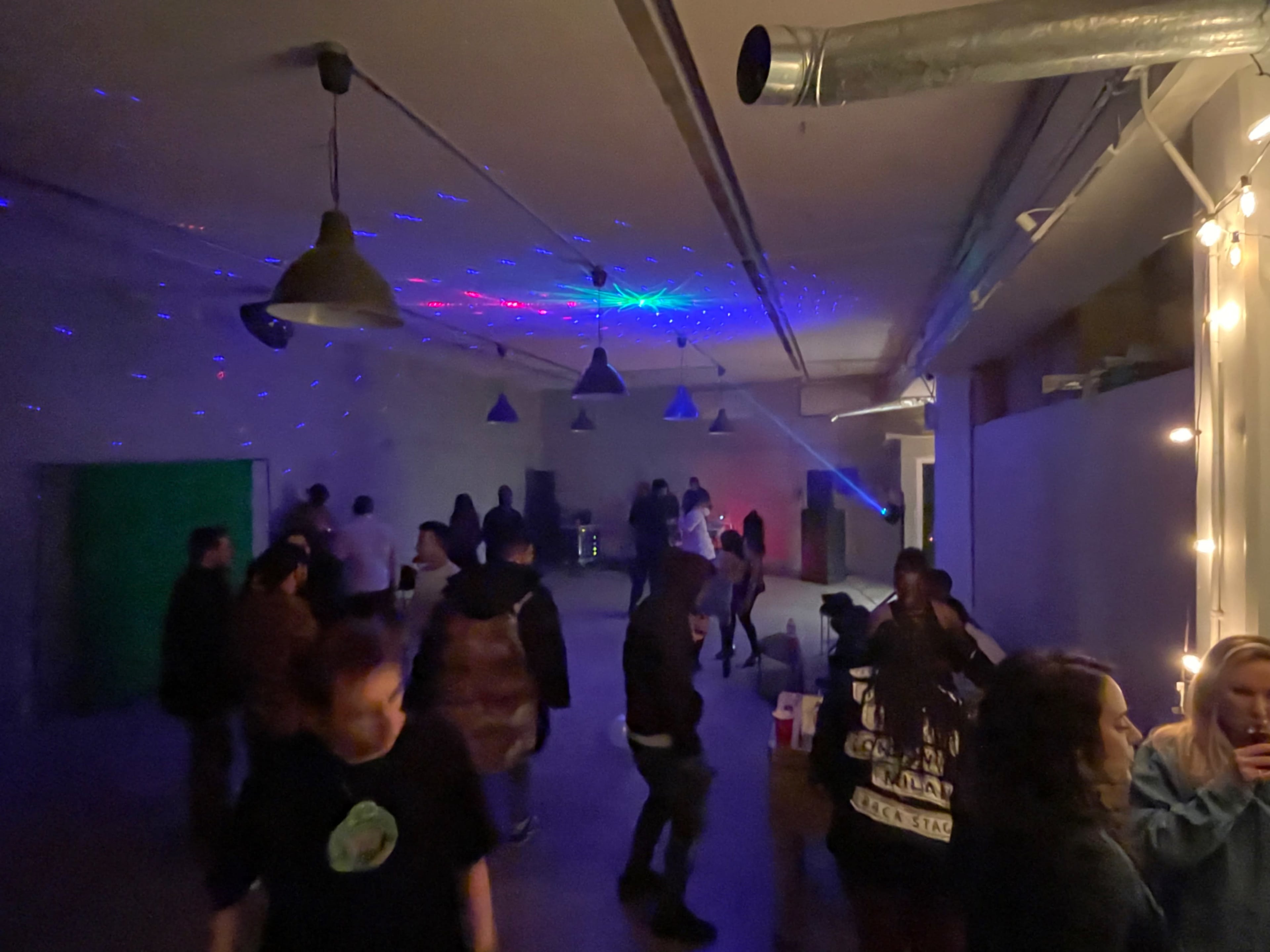 A group of people socialize and dance in a dimly lit room featuring colorful lights and a disco ball effect.