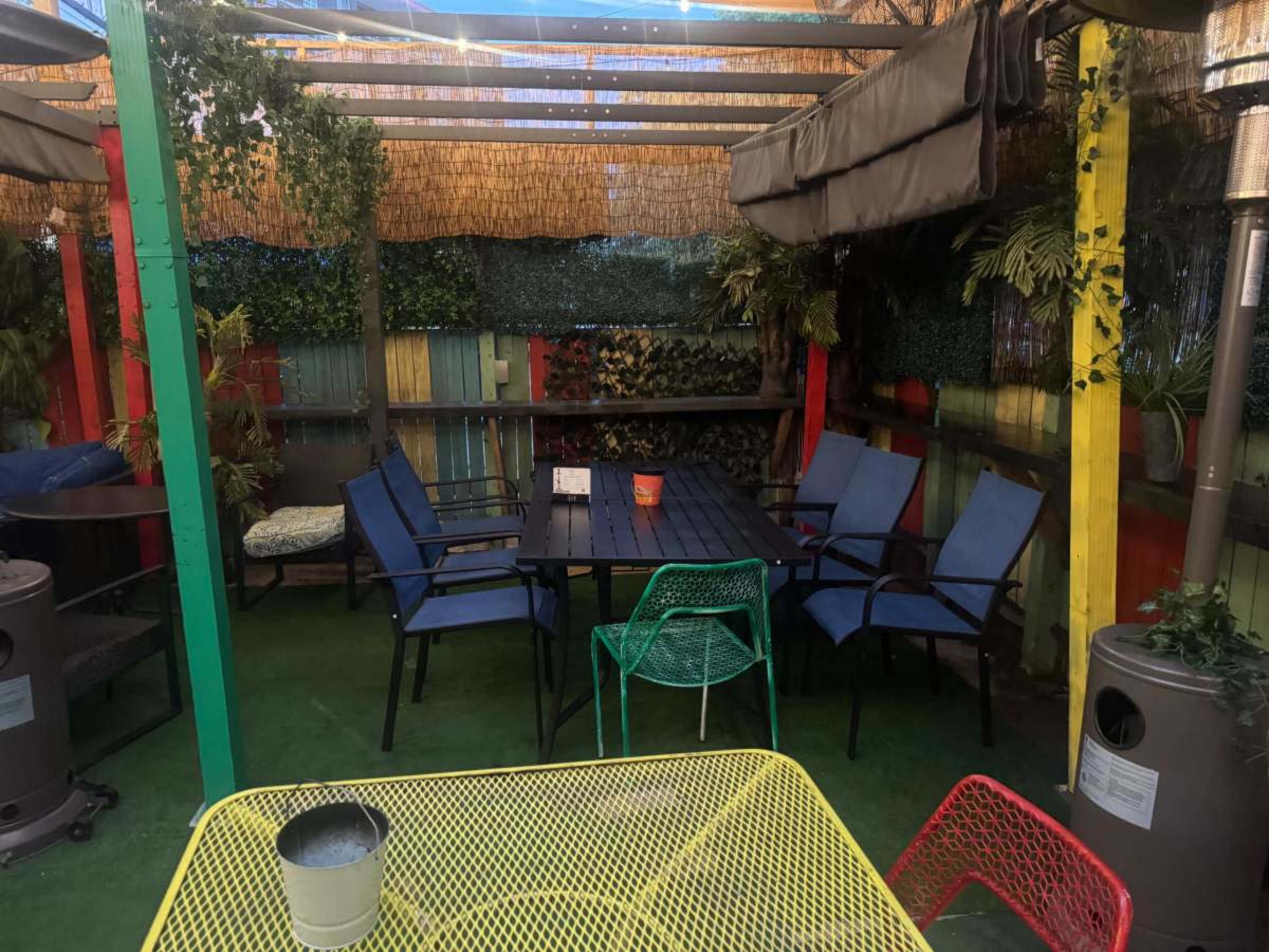 The image shows a colorful outdoor seating area with a large table surrounded by chairs, illuminated by overhead lighting and adorned with plants.
