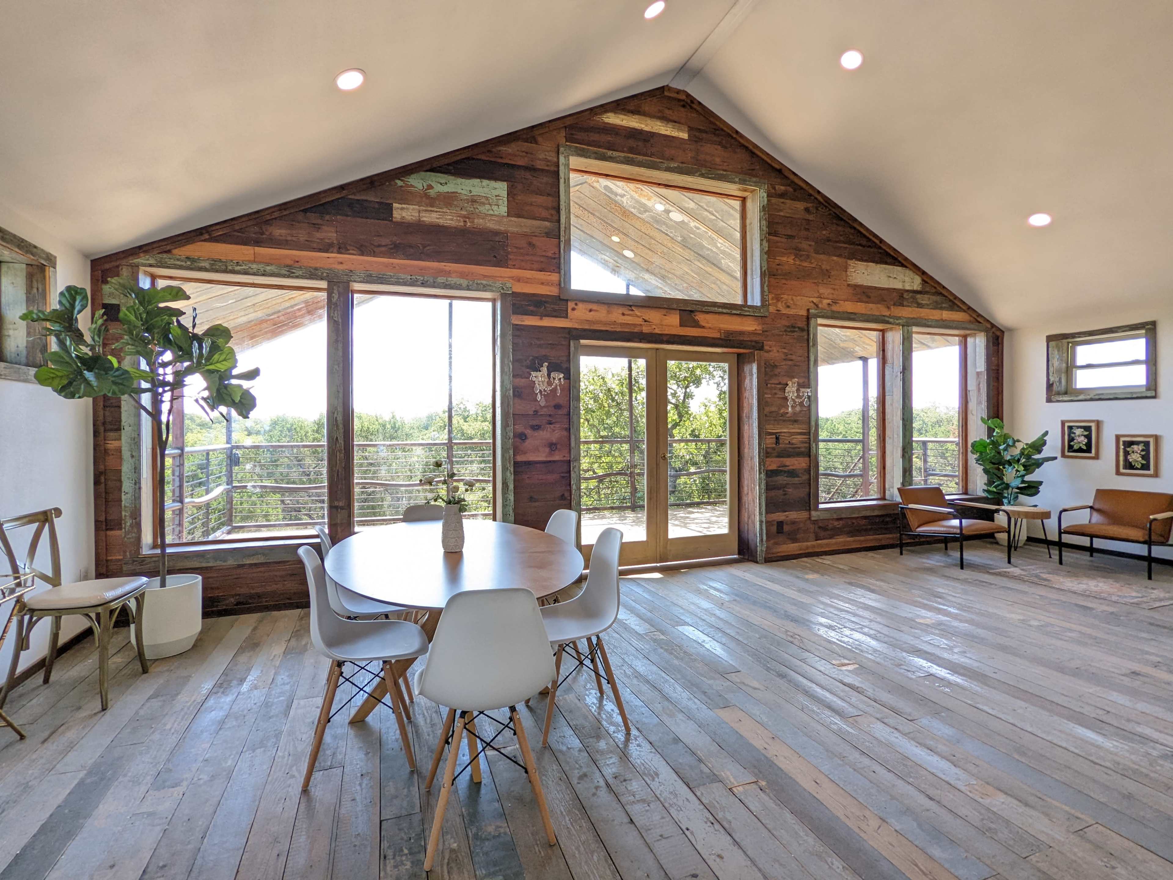 A spacious indoor area featuring a round dining table, modern chairs, large windows, and wooden walls, with a view of greenery outside.