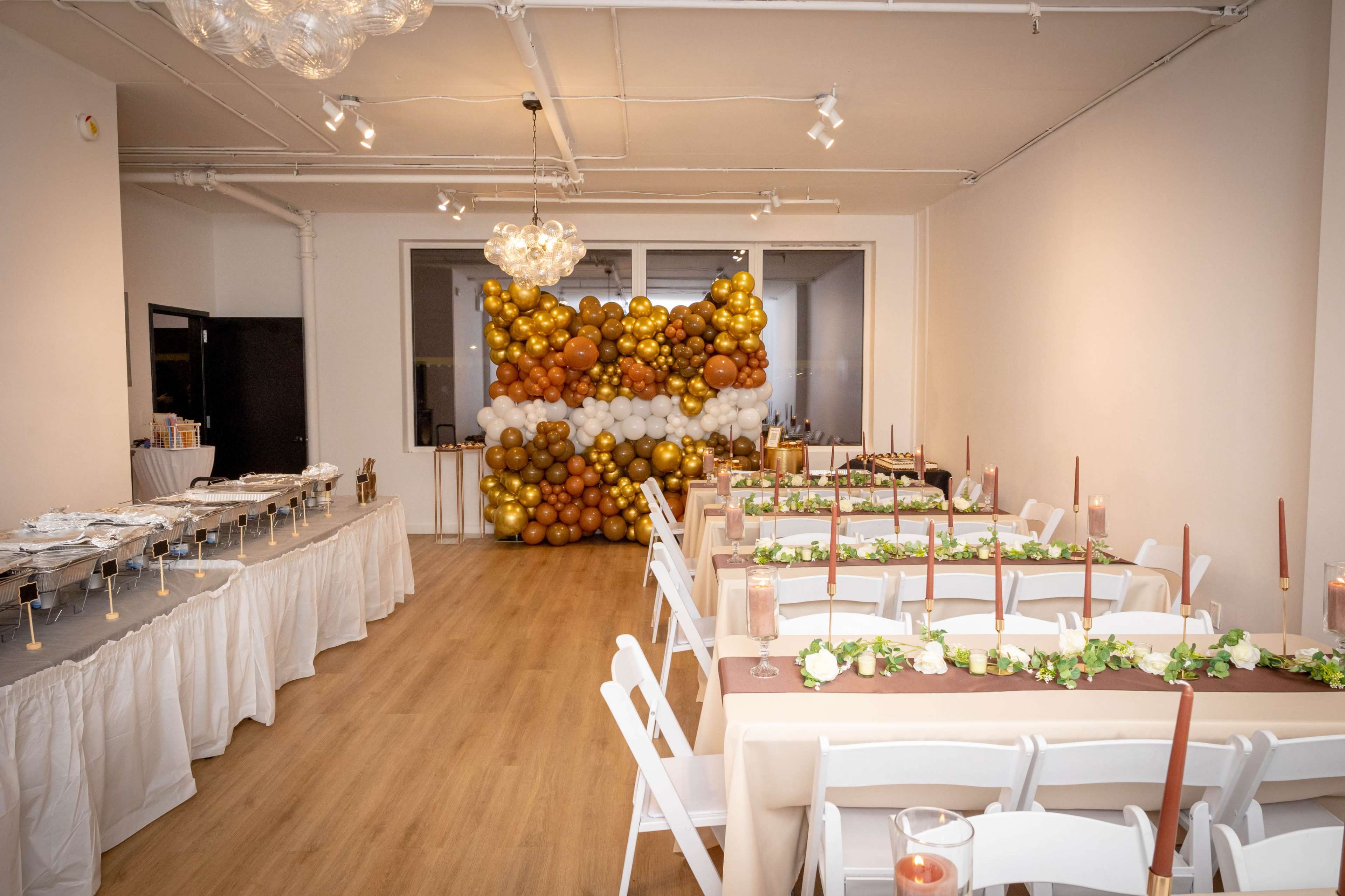 The image shows a banquet hall setup with long tables adorned with greenery and candles, and a decorative balloon backdrop in gold and white.