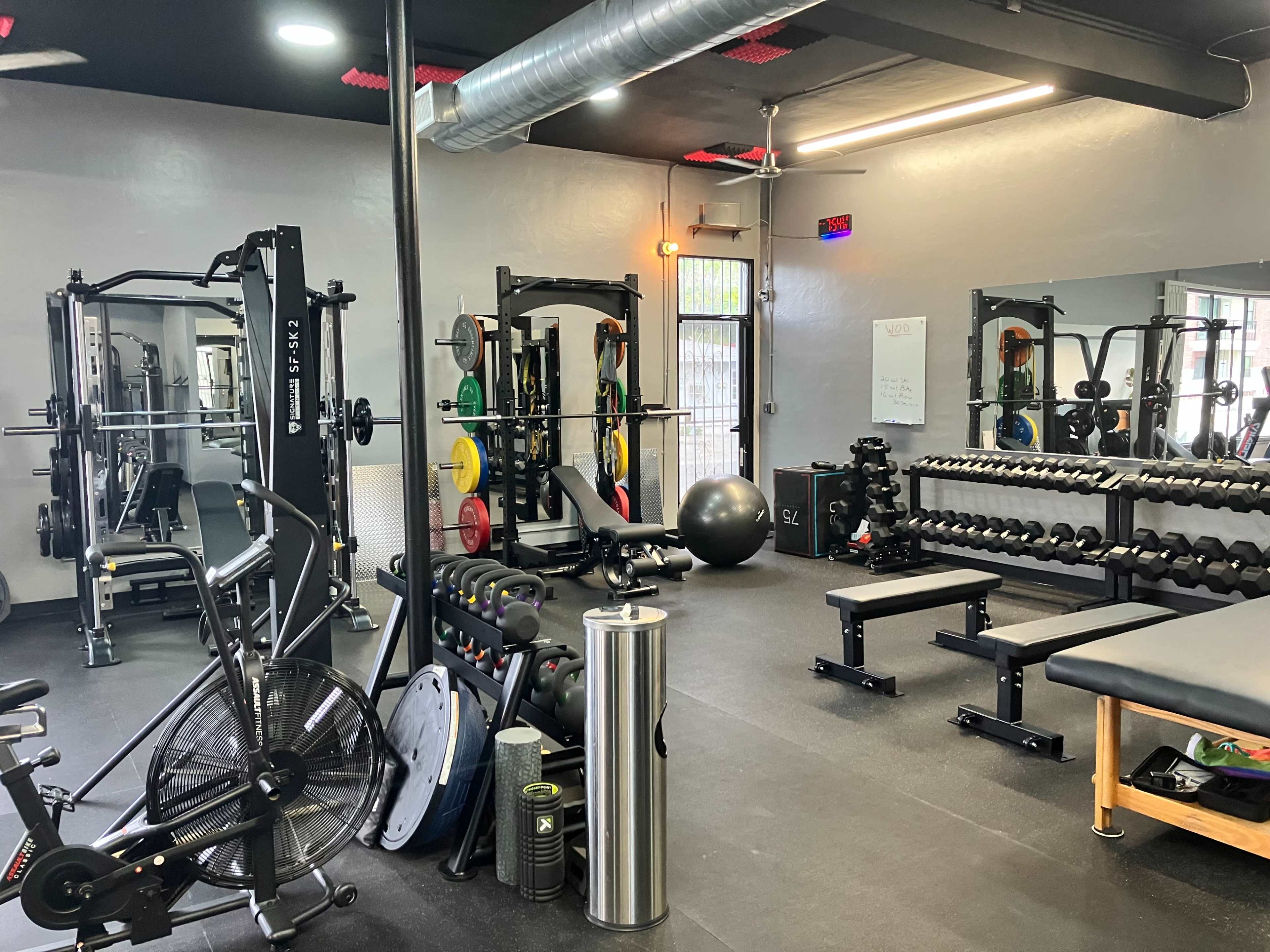 The image shows a well-equipped gym interior featuring various weightlifting machines, free weights, and fitness accessories.