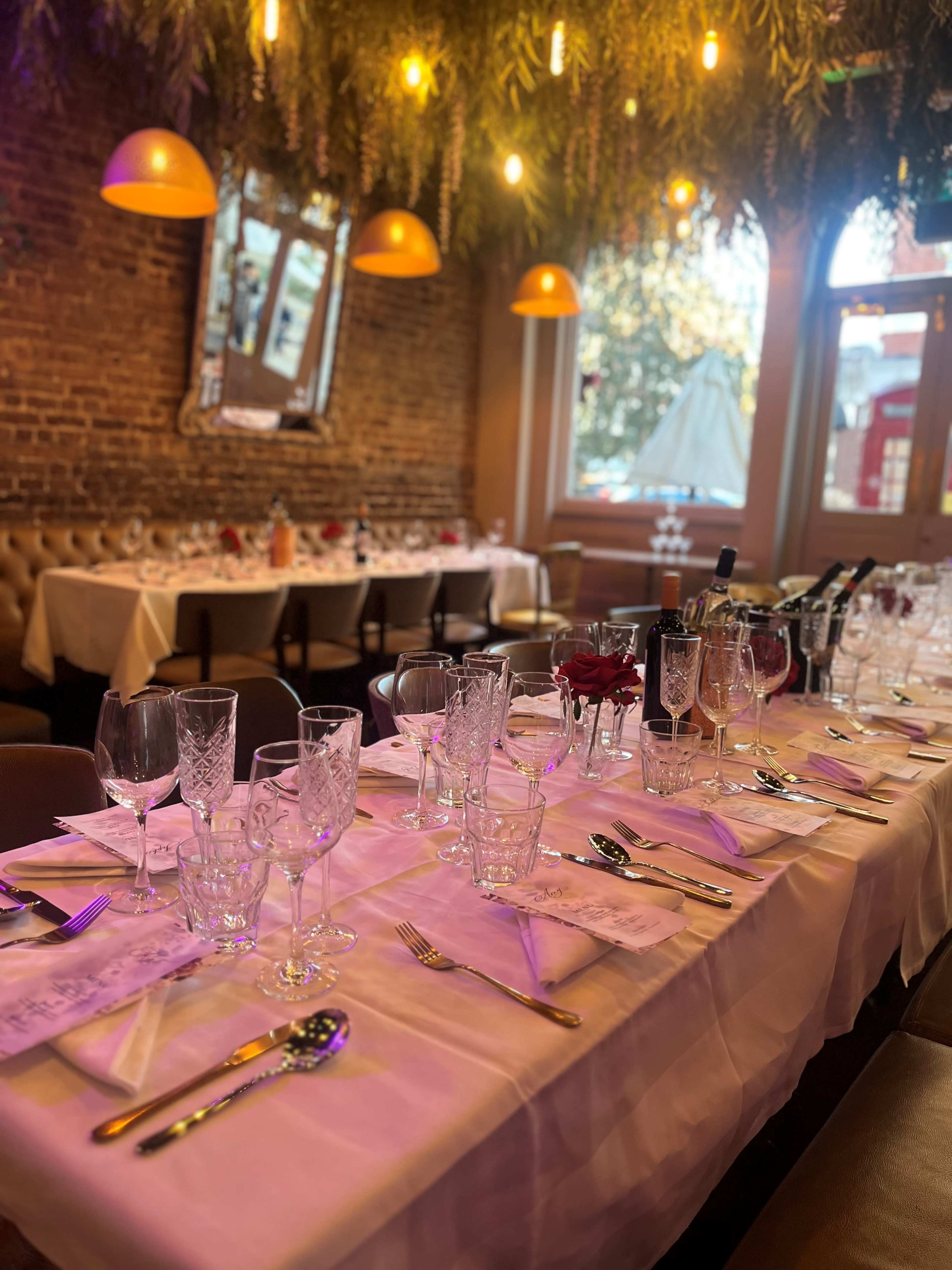 A long dining table is set with decorative glassware, silverware, and floral arrangements, under warm hanging lights and greenery.