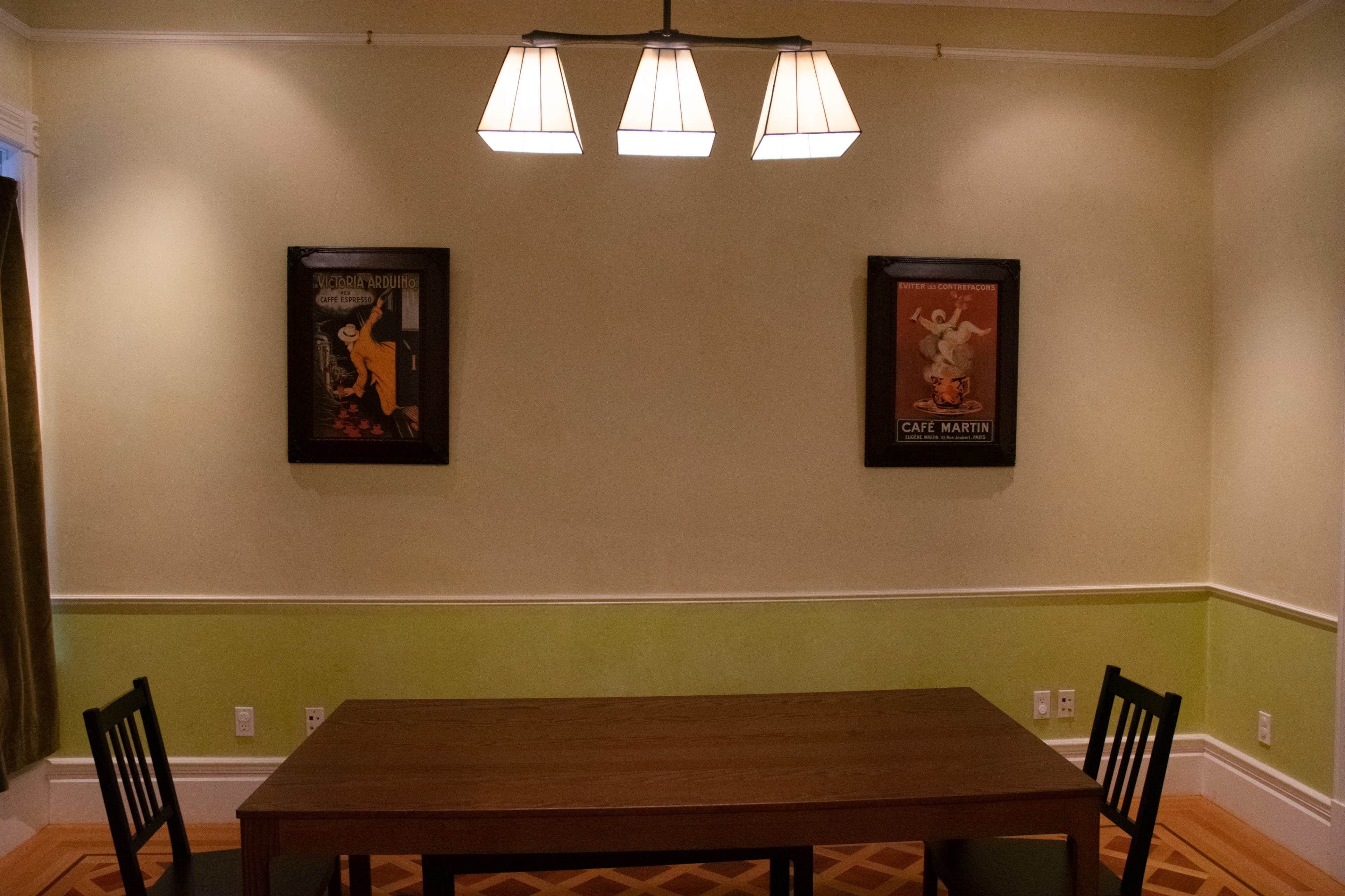 A simple dining room features a wooden table with two black chairs, surrounded by light-colored walls and framed vintage posters on either side.