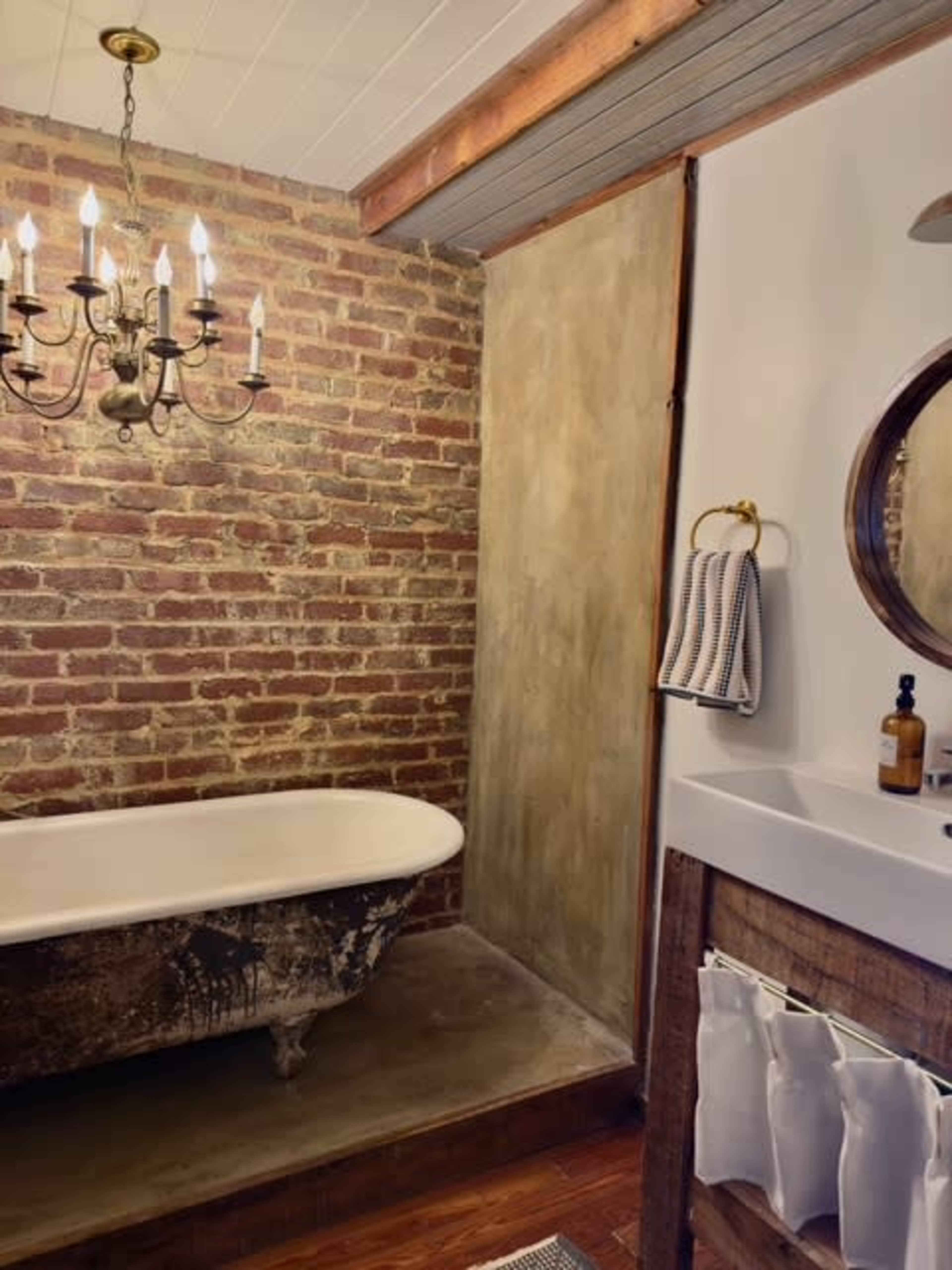 The image shows a bathroom featuring a vintage clawfoot tub, exposed brick wall, and a wooden sink cabinet with a round mirror above it.