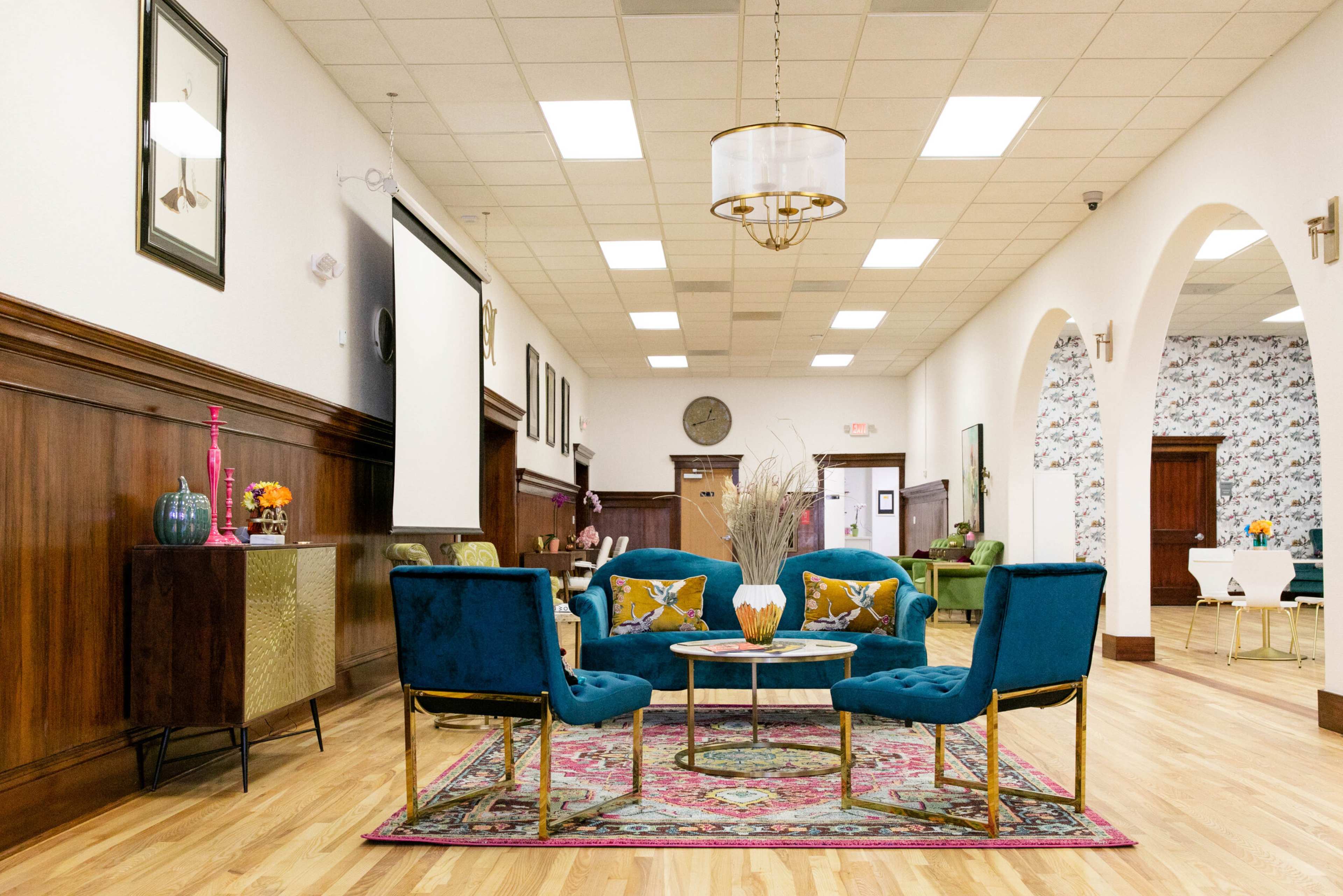 A spacious interior with wooden paneling, featuring a seating area with four blue chairs around a coffee table on a patterned rug, along with decorative furnishings and wall art.