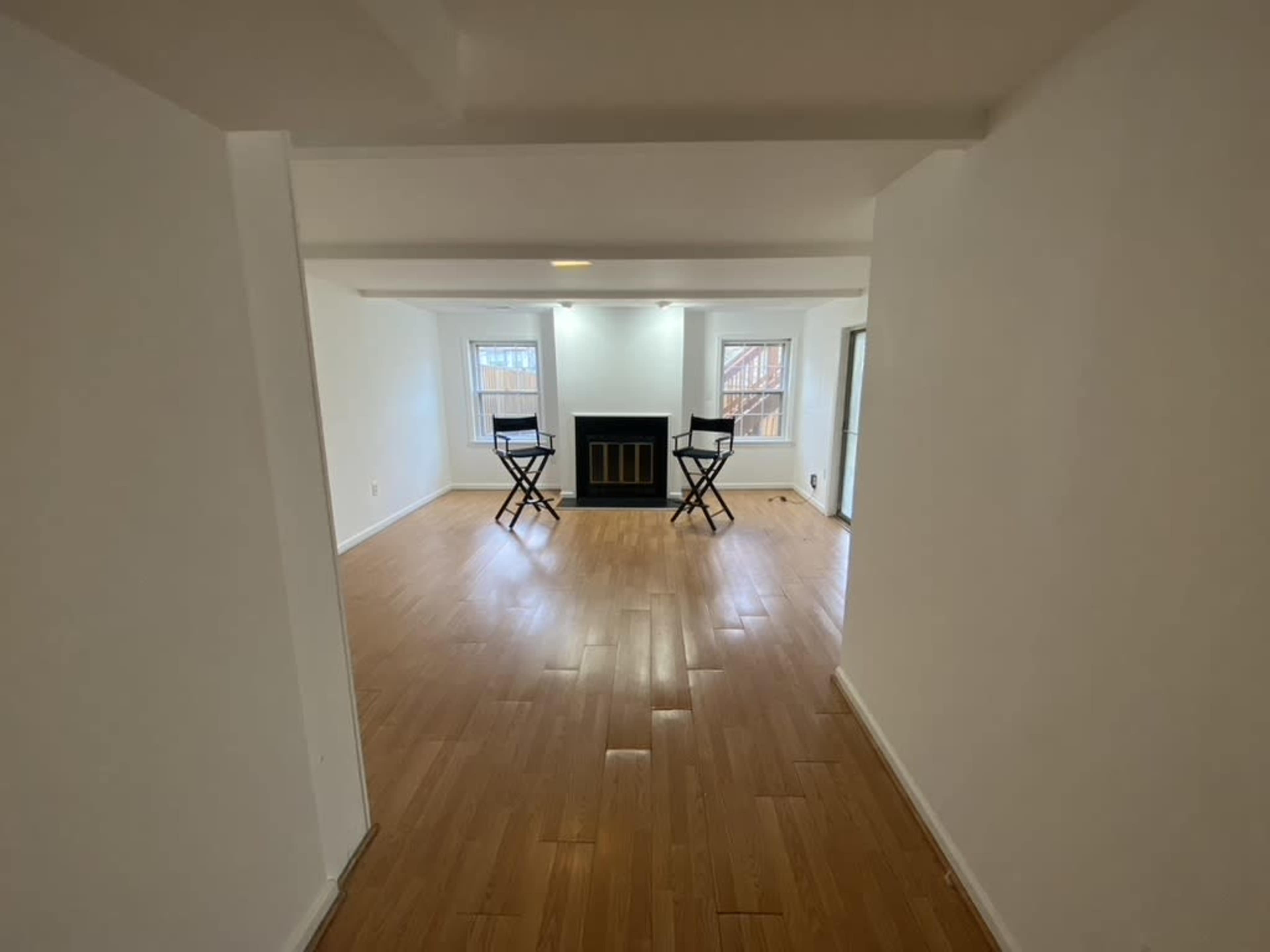 A spacious, empty room with wooden flooring, two black stools facing a fireplace, and large windows letting in natural light.