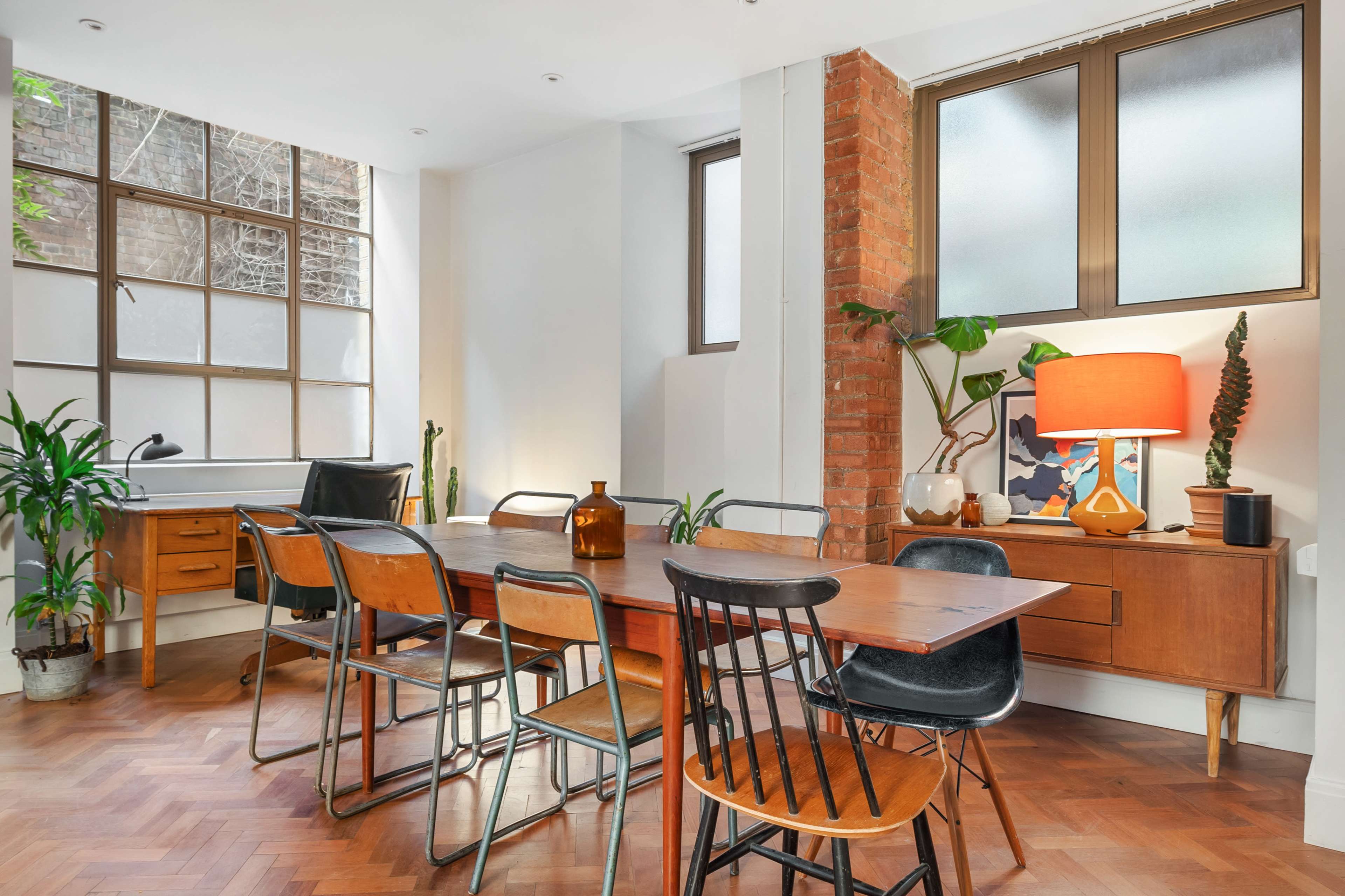 The image shows a modern dining area with a wooden table surrounded by various chairs, plants in the corners, and large windows allowing natural light to enter.